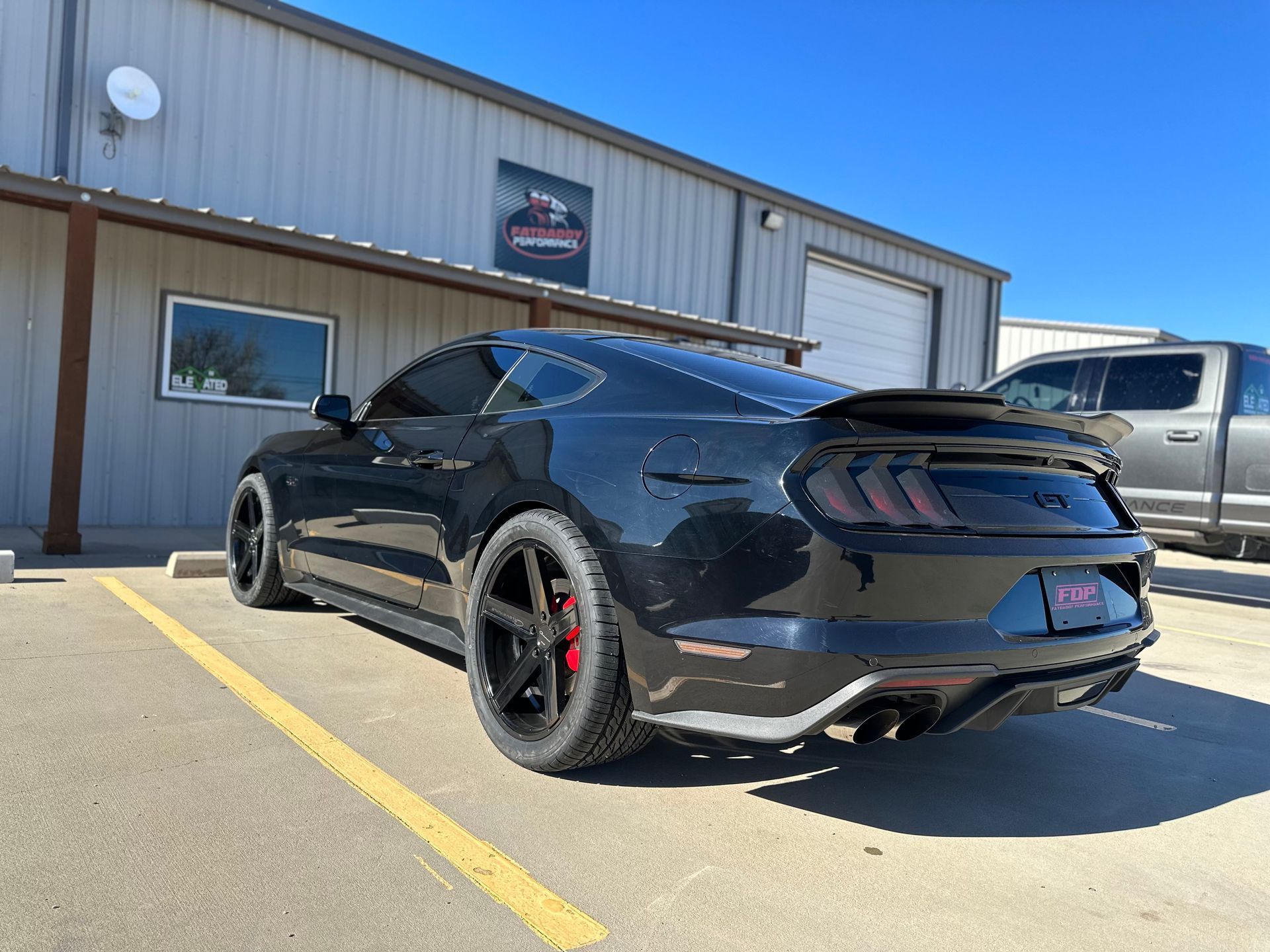 Black Ford Mustang parked in front of a building with a gray truck in the background.