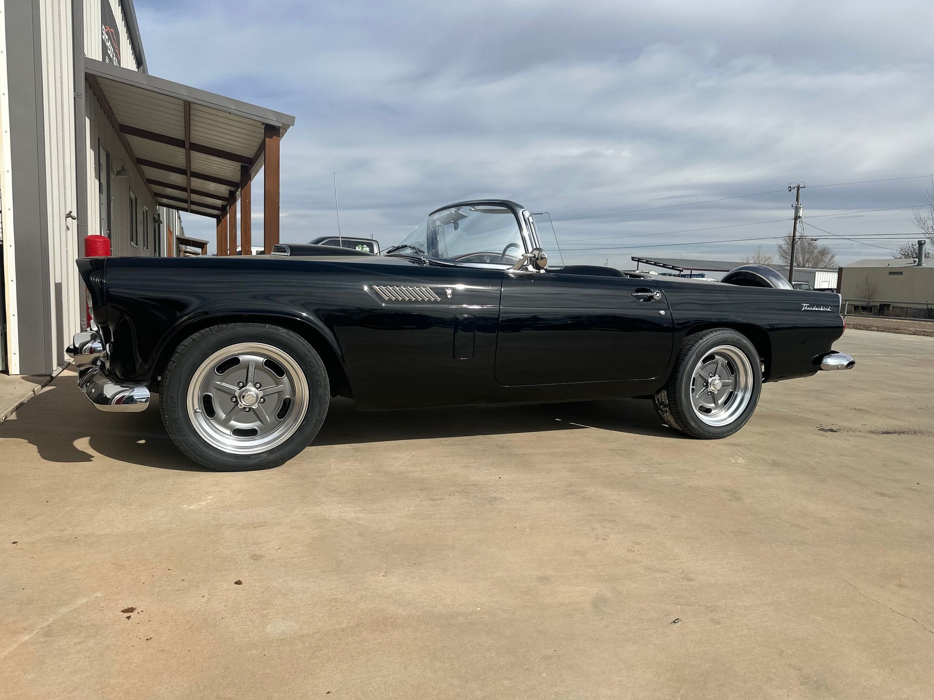 Black convertible car parked in front of a building on a concrete surface under a cloudy sky.