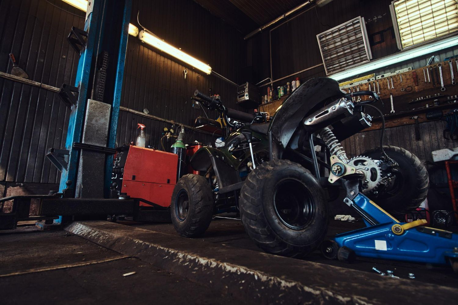 ATV in a dimly lit garage, raised on a jack, with tools and equipment nearby.