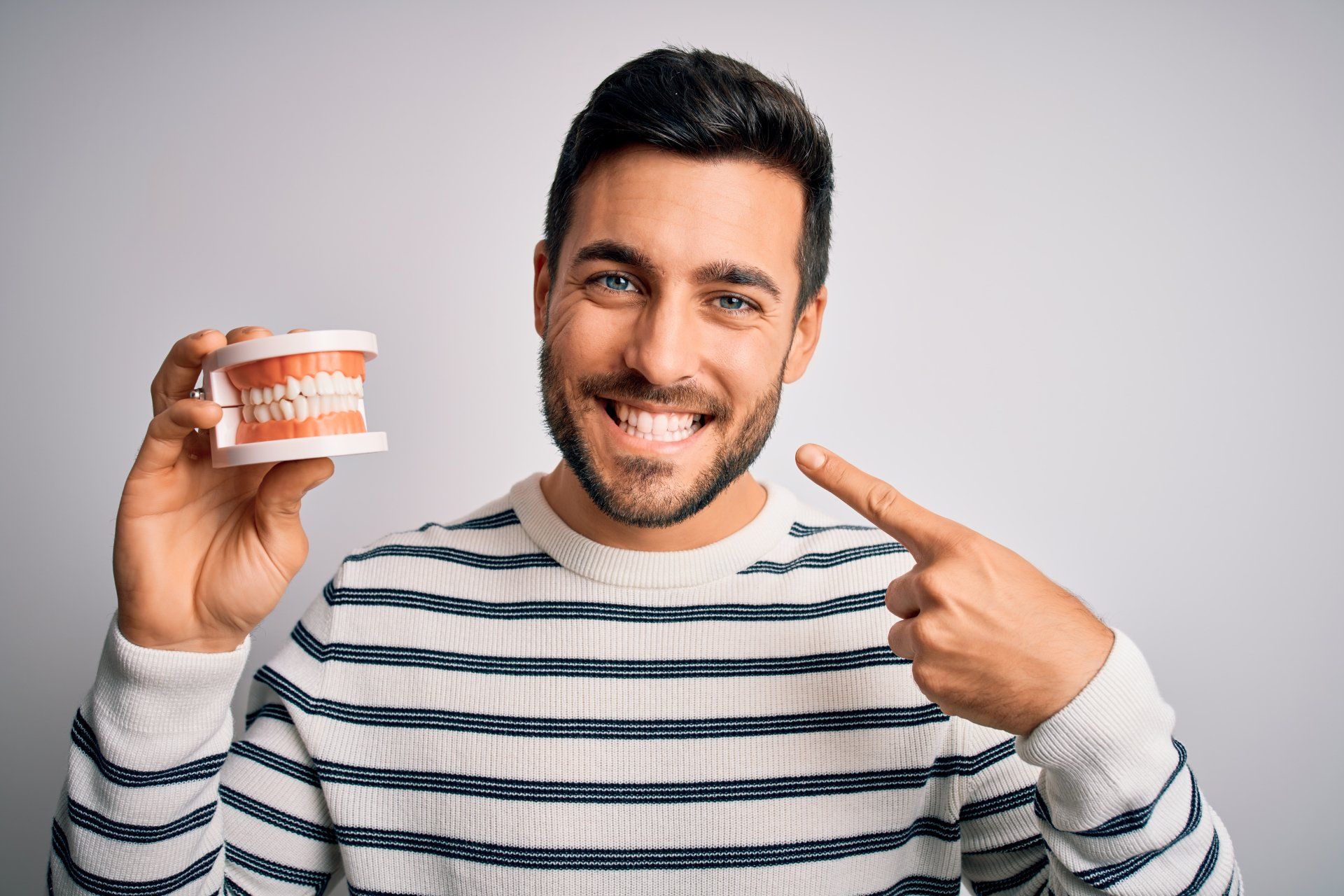 Implant supported dentures example guy smiling a happy smile using one hand to point to his mouth and another to hold a model of upper and lower teeth