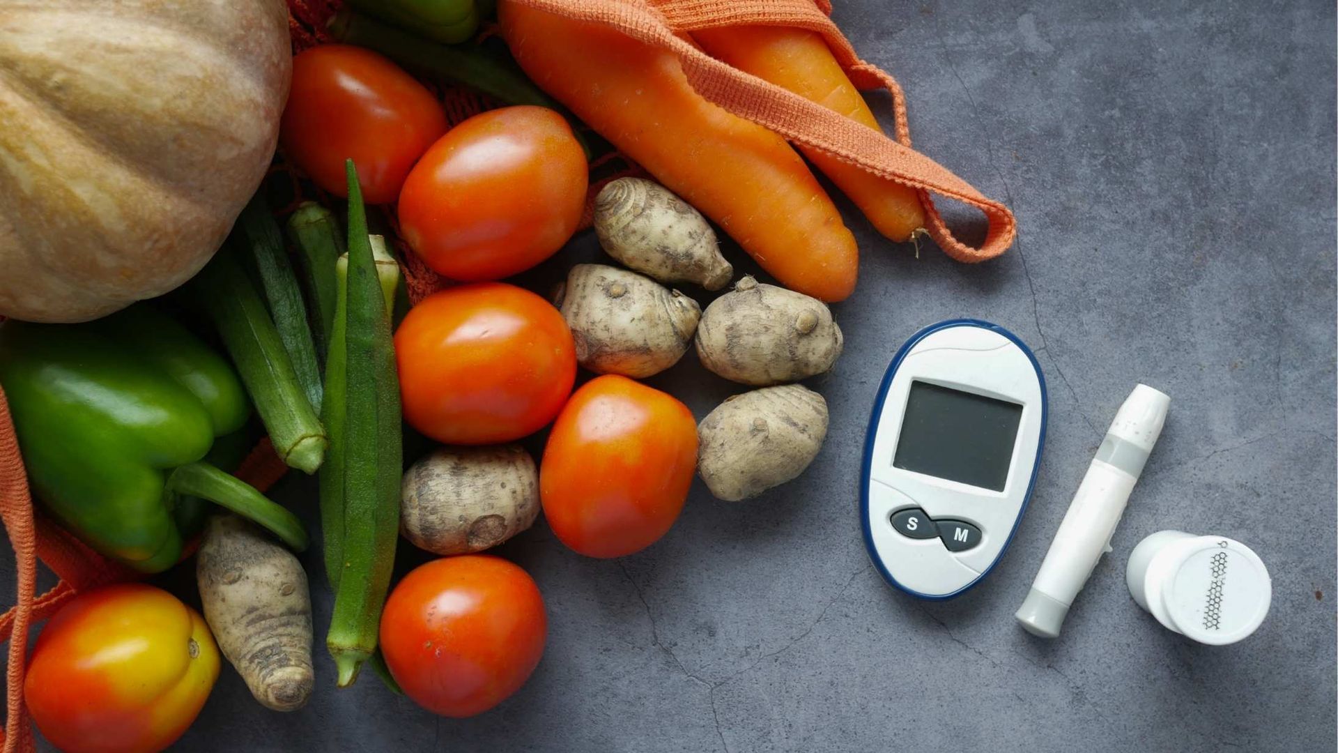 Assortment of vegetables spilling from a reusable bag next to a blood glucose meter and testing supplies on a gray surface.
