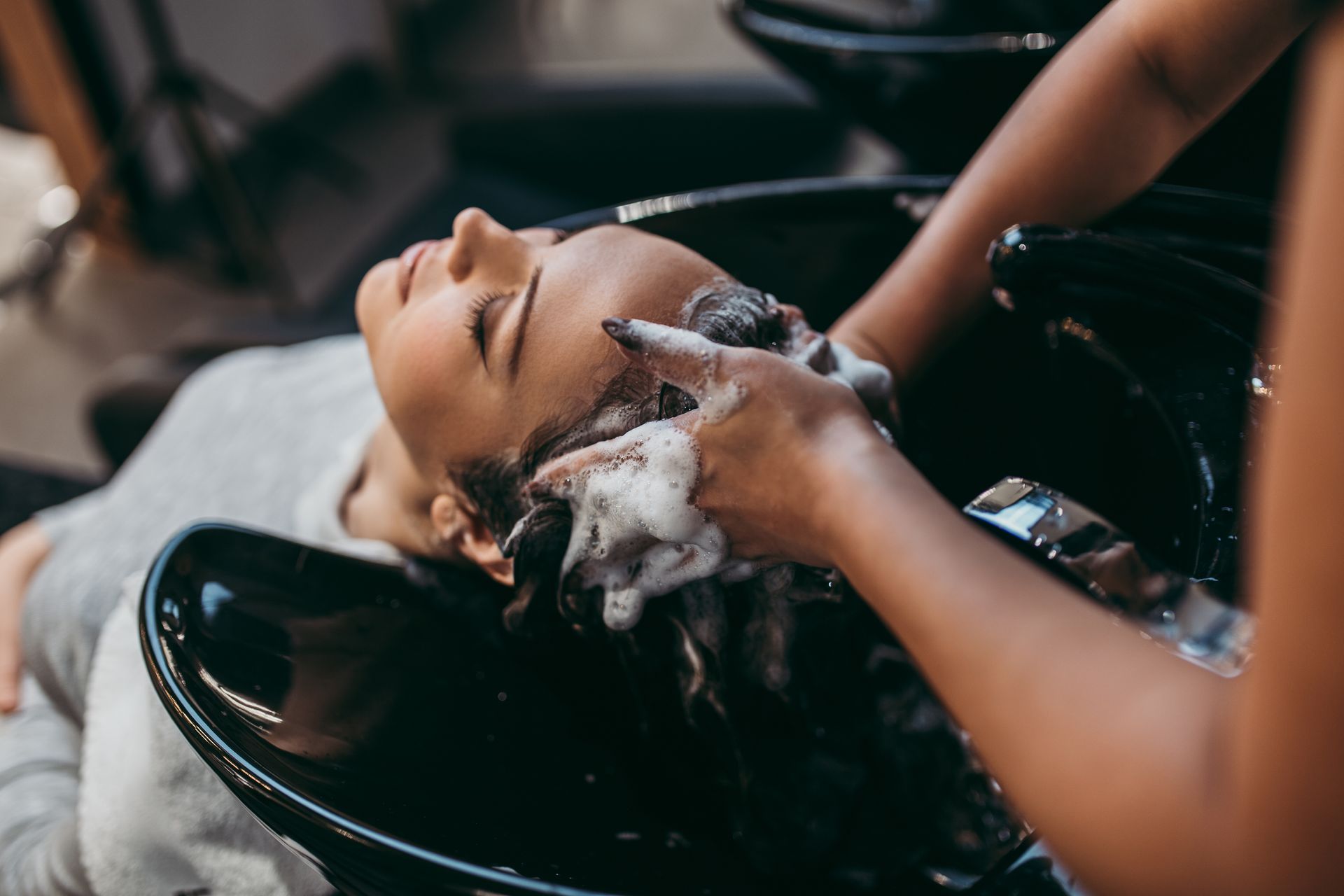 Person having hair washed at a salon, black bowl, shampoo foam, hands, closed eyes.
