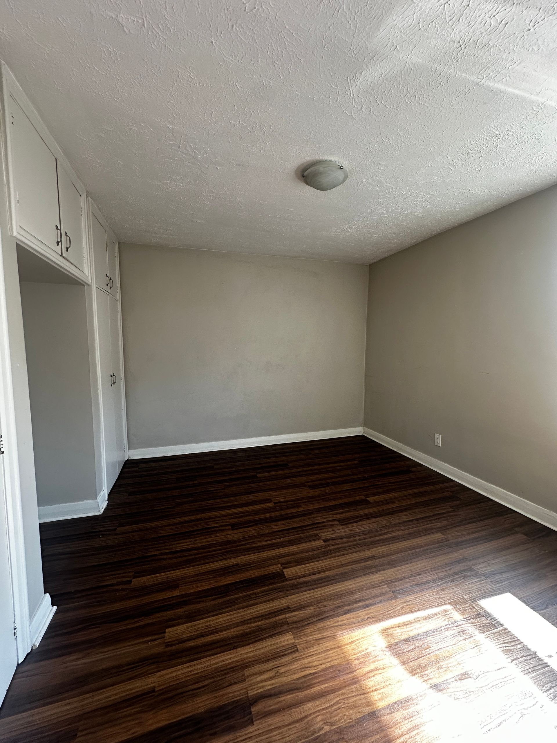 Empty unfurnished bedroom with beige walls, dark wood floor, closet, and bright sunlight on the floor
