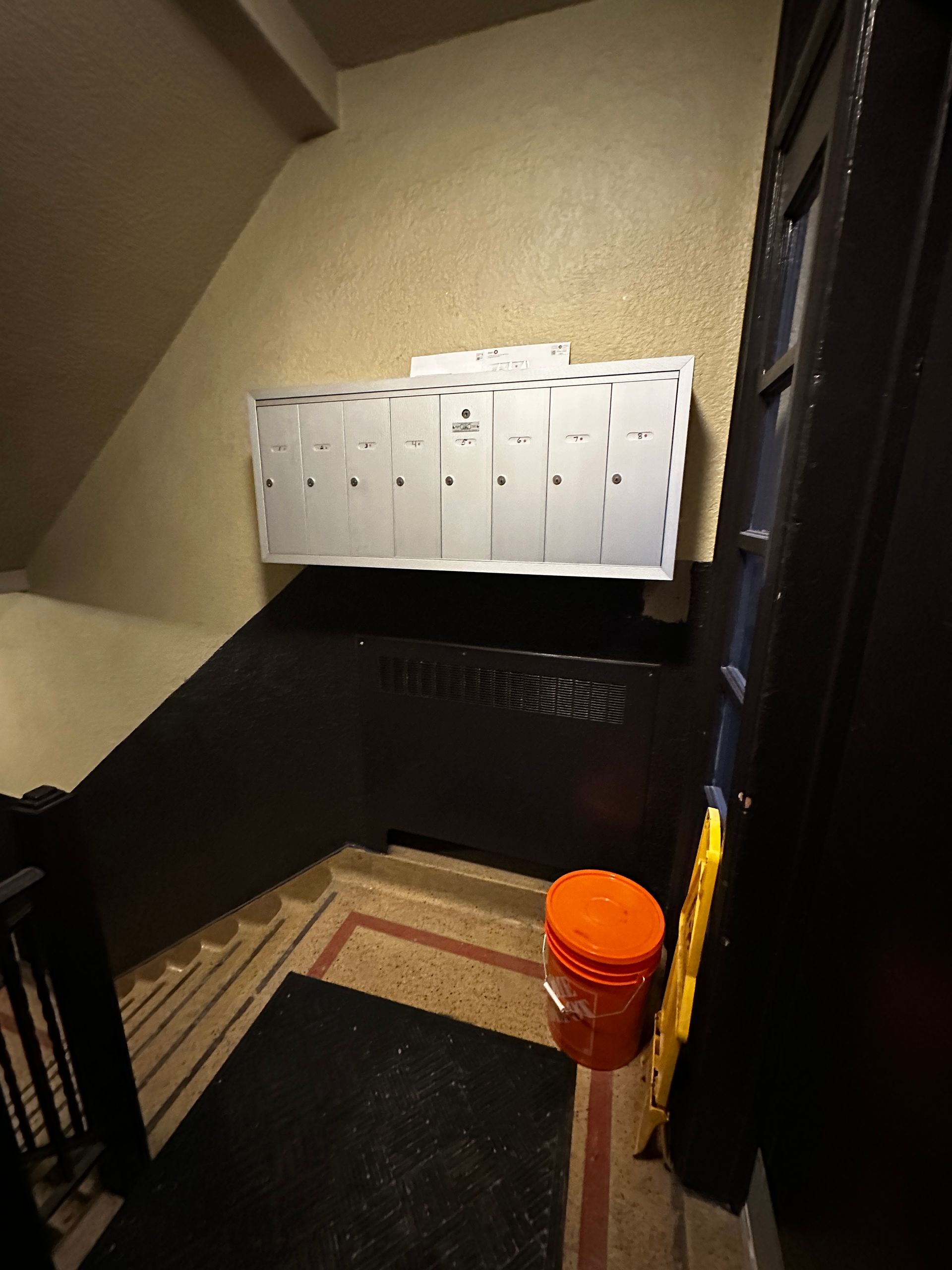 Stairwell landing with a wall of white mailboxes and an orange bucket on the floor.