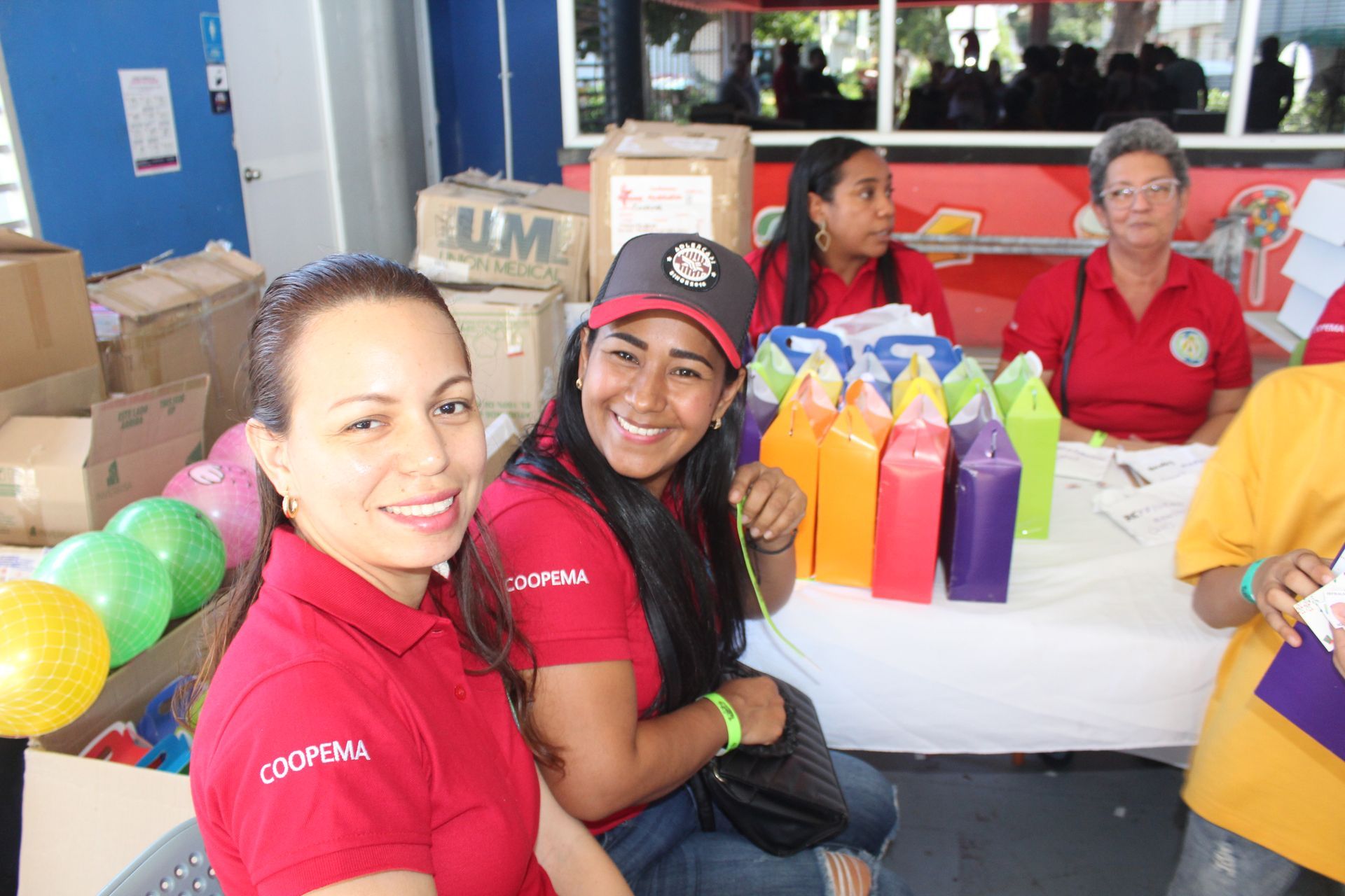 Volunteers in red shirts smiling at a table with colorful bottles and balloons at an indoor event