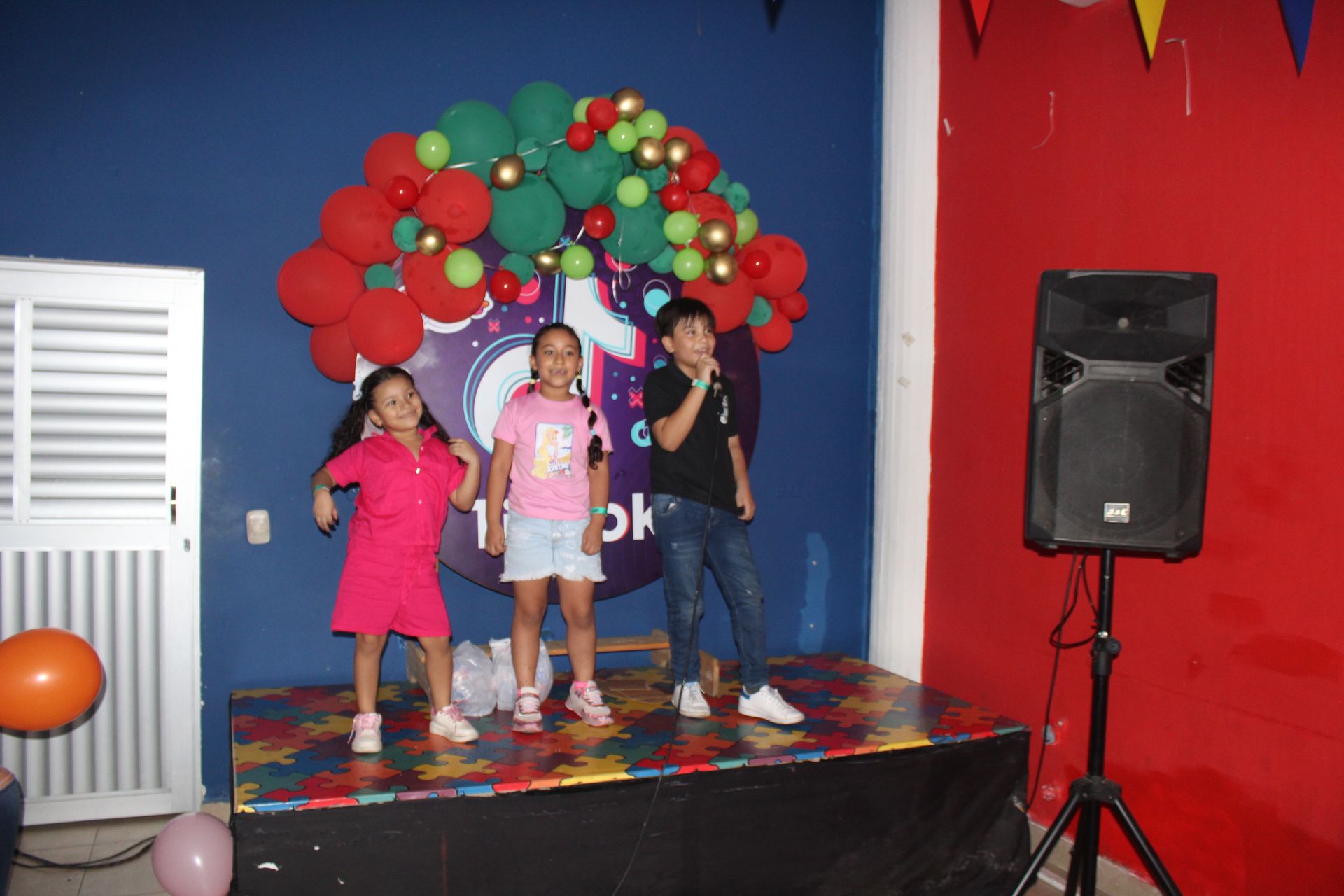 Three children posing on a small stage with a red and green balloon arch and a speaker beside them.