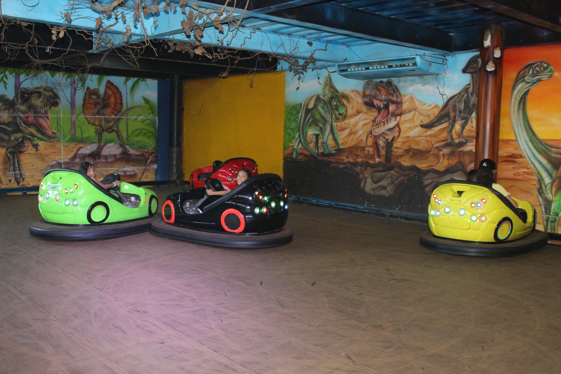 Three bumper cars on an indoor ride, including green, black-red, and yellow cars under a painted ceiling.