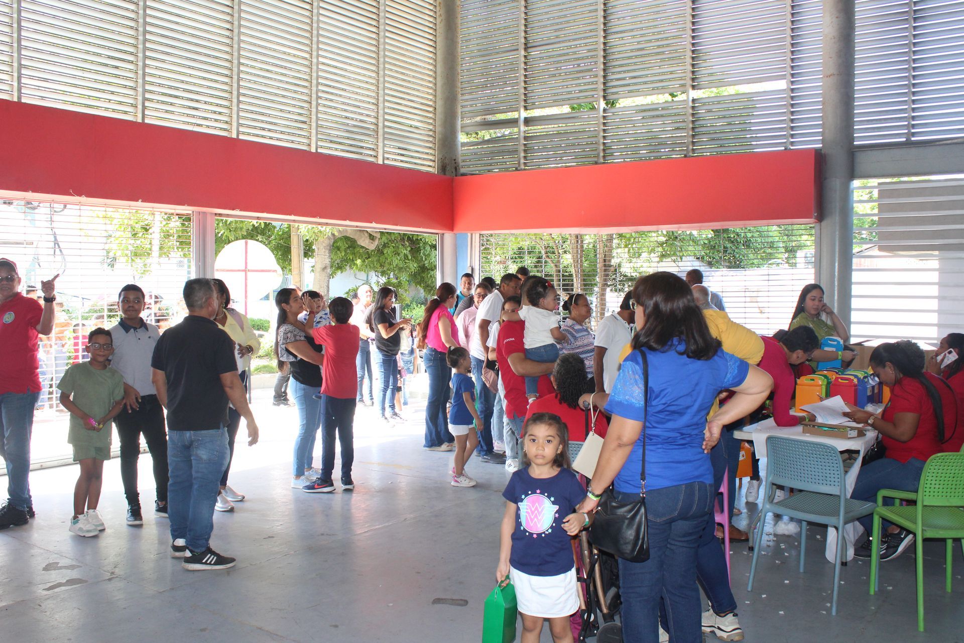 People gathered in a bright indoor hall, with a woman and child in the foreground and others seated and standing nearby.