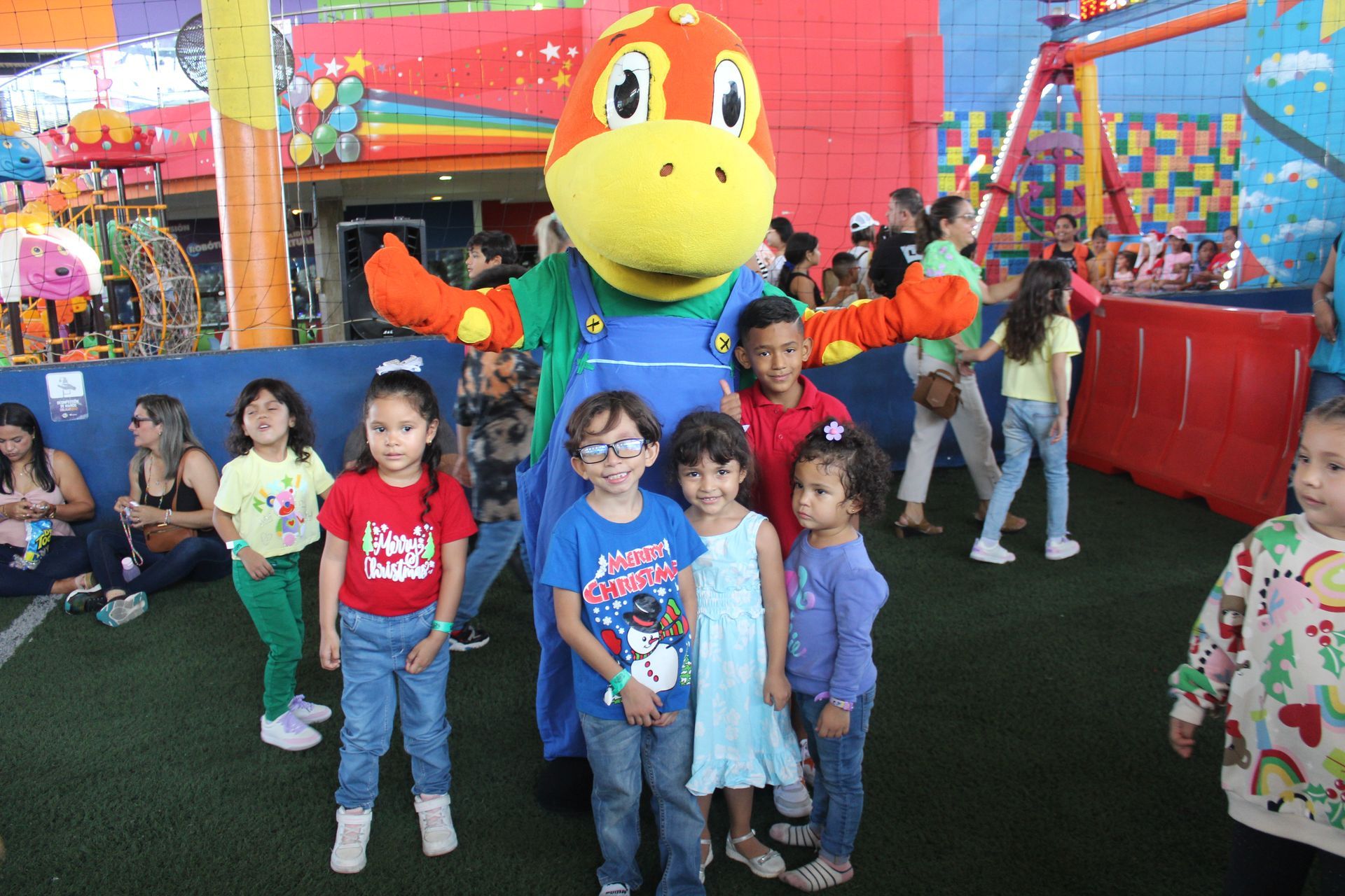 Children posing with a large colorful cartoon mascot at an indoor playground, with bright play structures in the background