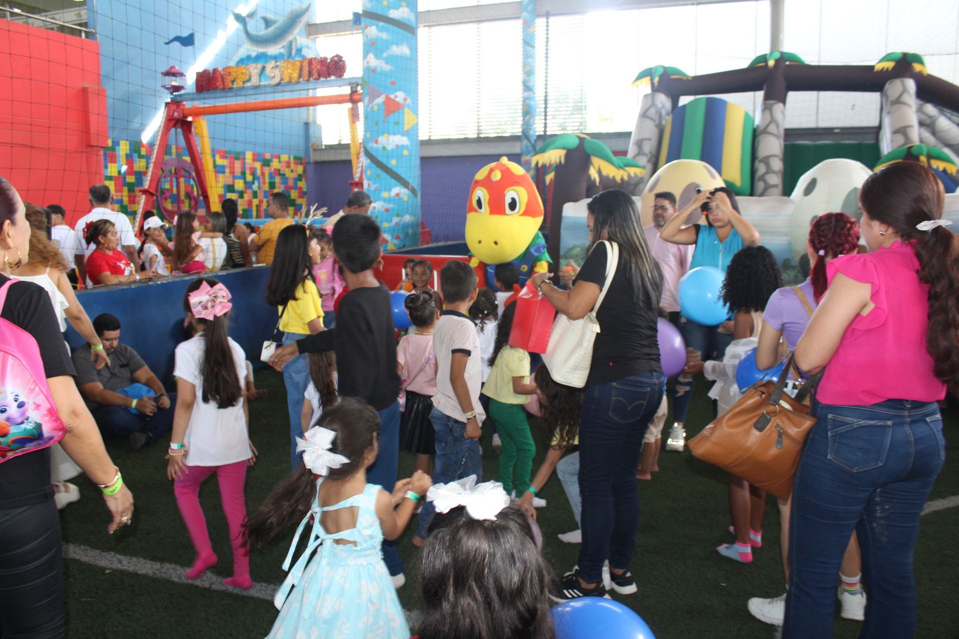 Crowded outdoor children’s party with a colorful bird mascot, bounce house, and kids dancing on grass