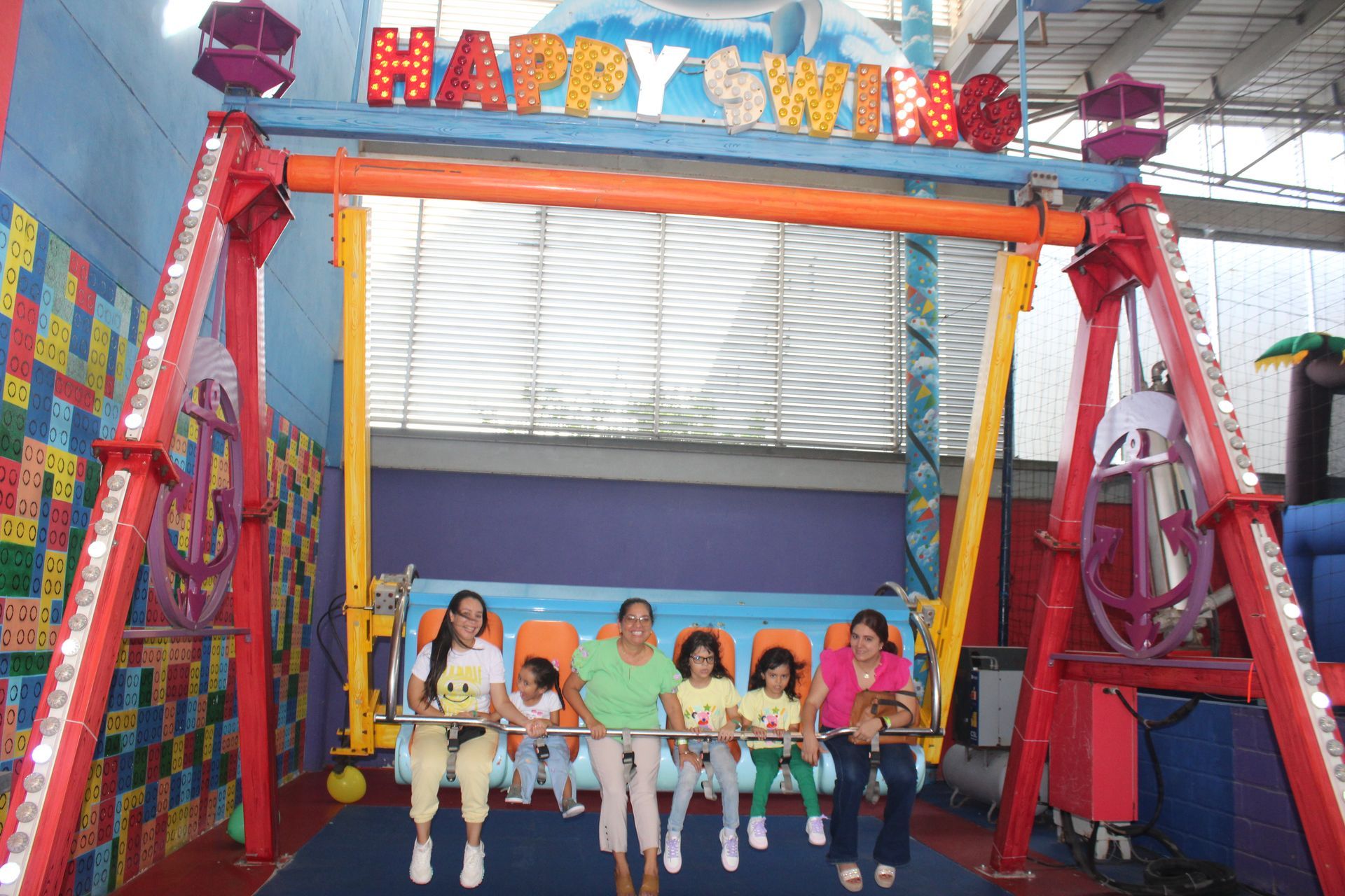 People seated on a bright amusement ride named “HAPPY SWING” inside a covered structure.