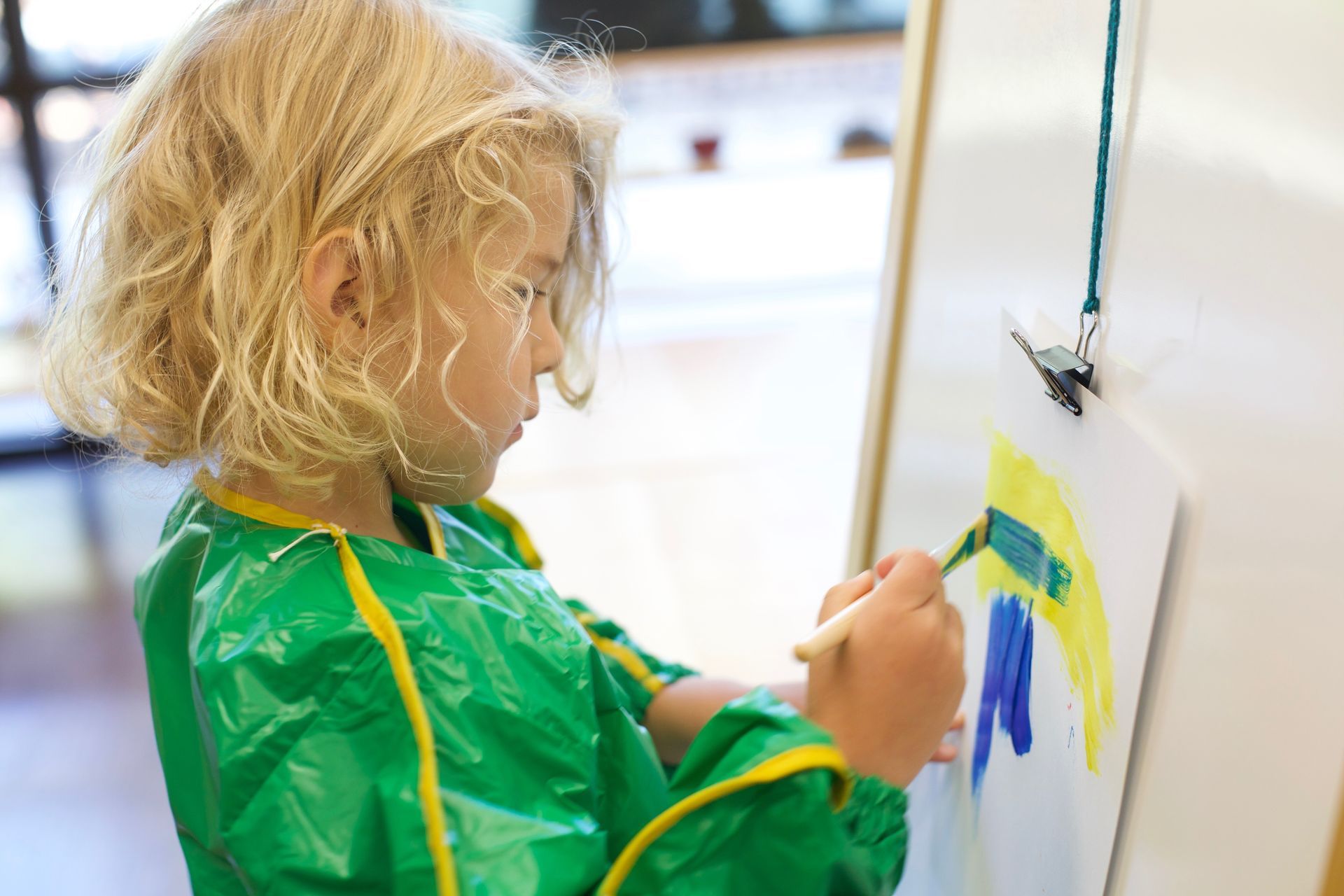 Child painting at an easel, wearing a green smock with yellow trim, in a bright art class