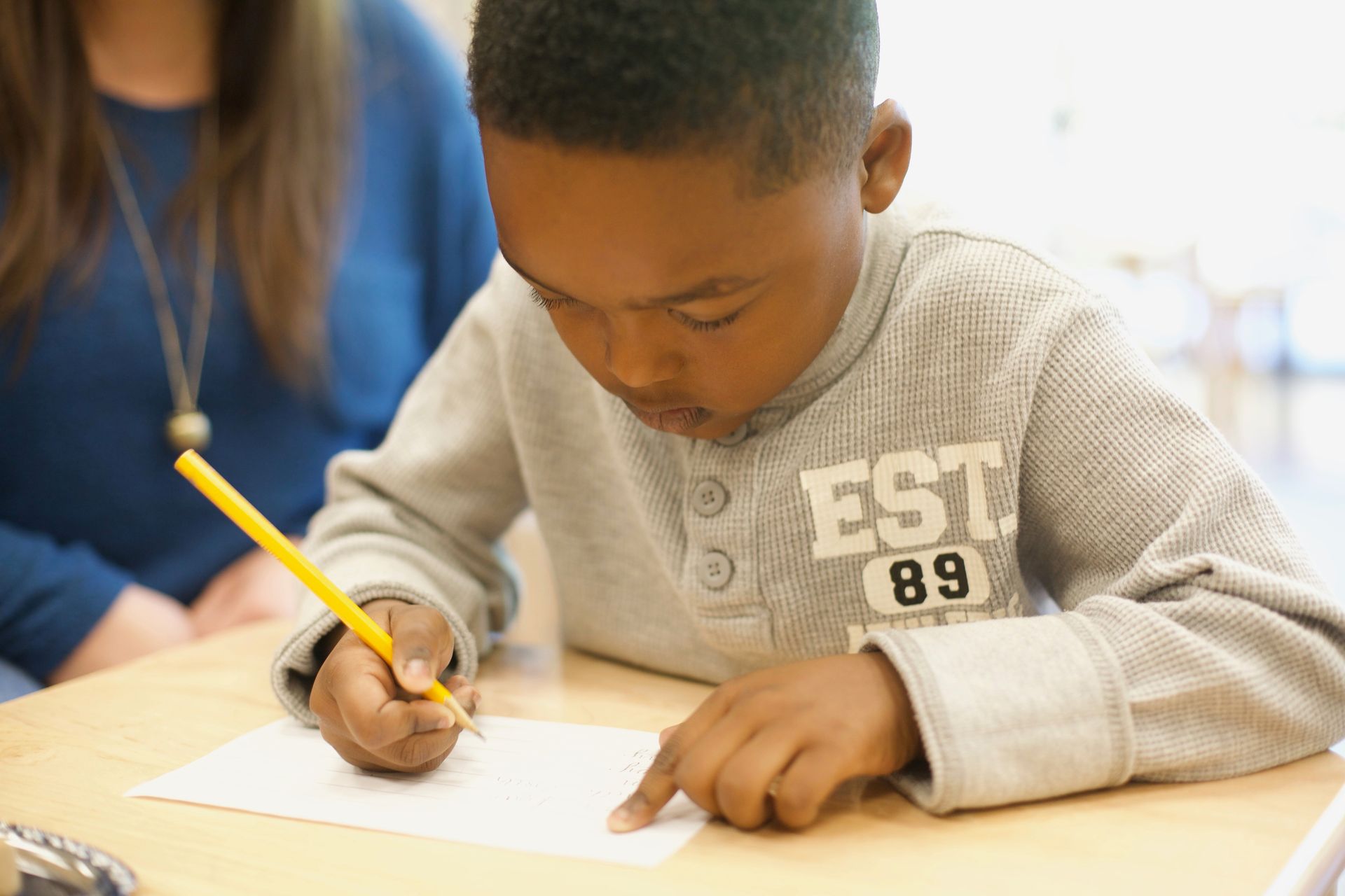 Child writing with a pencil at a desk in a classroom, with an adult nearby.