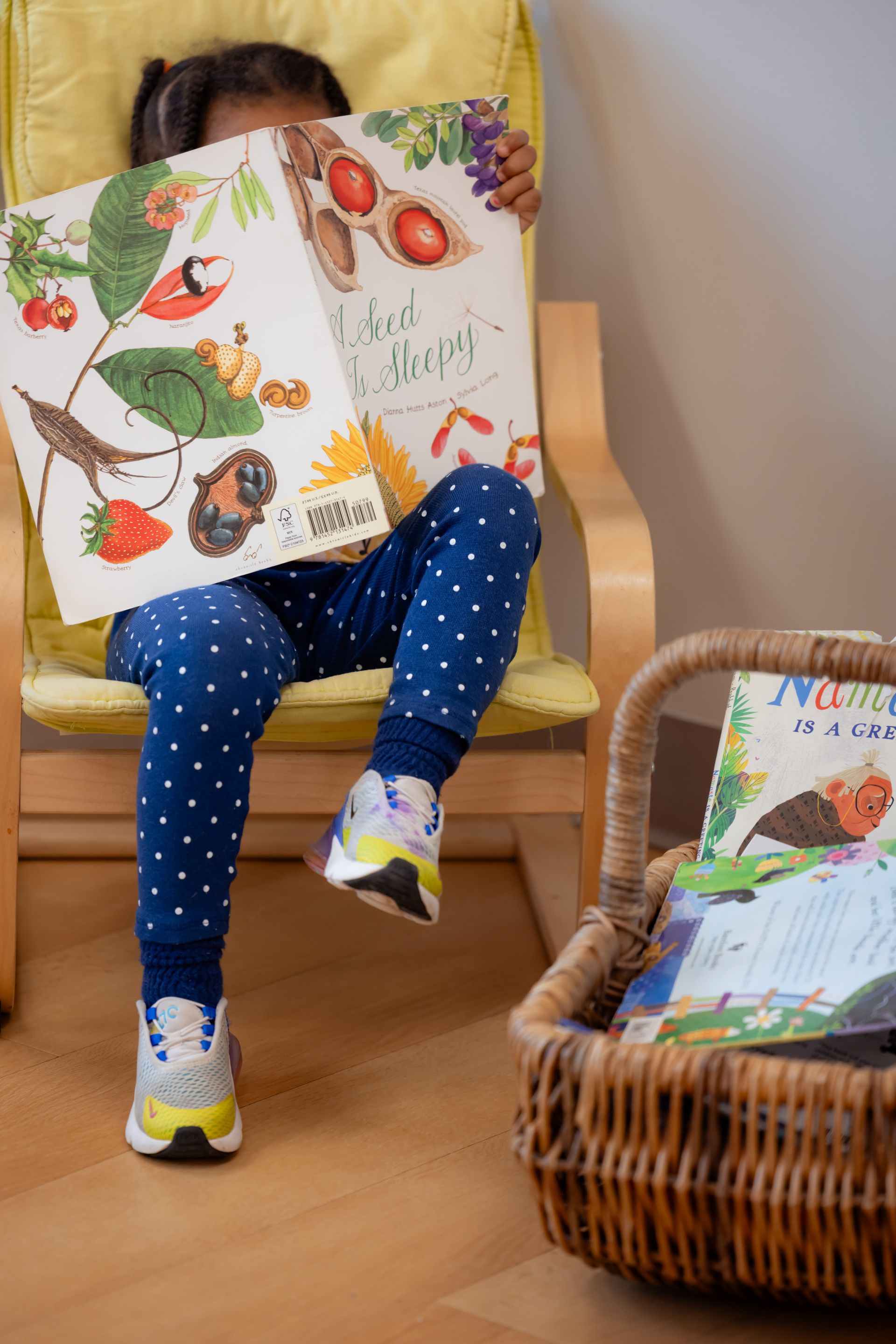 Child sitting in a yellow chair, reading a colorful picture book with a wicker basket of books beside them.