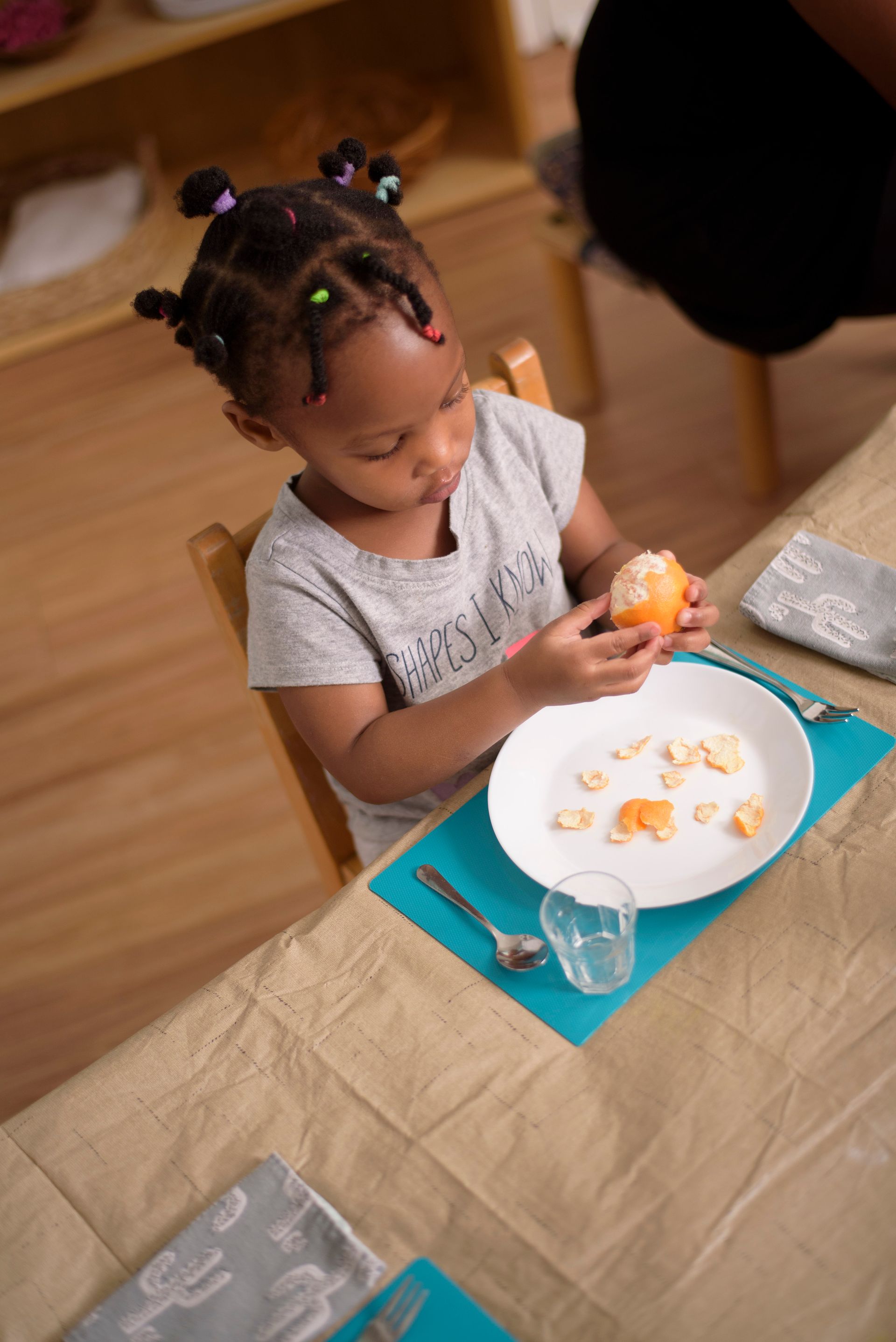 Child peeling an orange at a dining table with a plate, glass, and teal placemat.