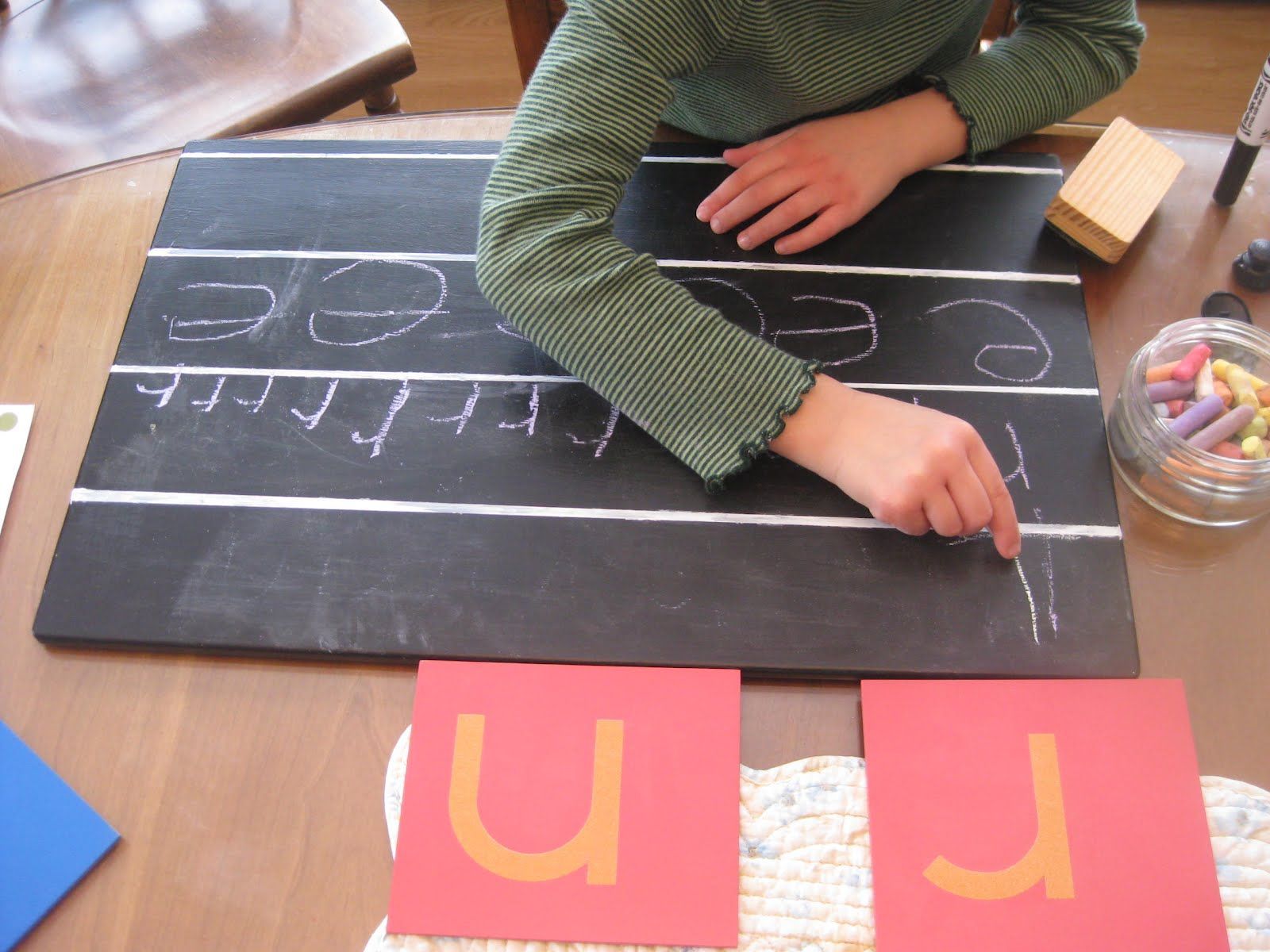 Montessori child writing letters on a chalkboard, with red letter cards on a table in front of them.
