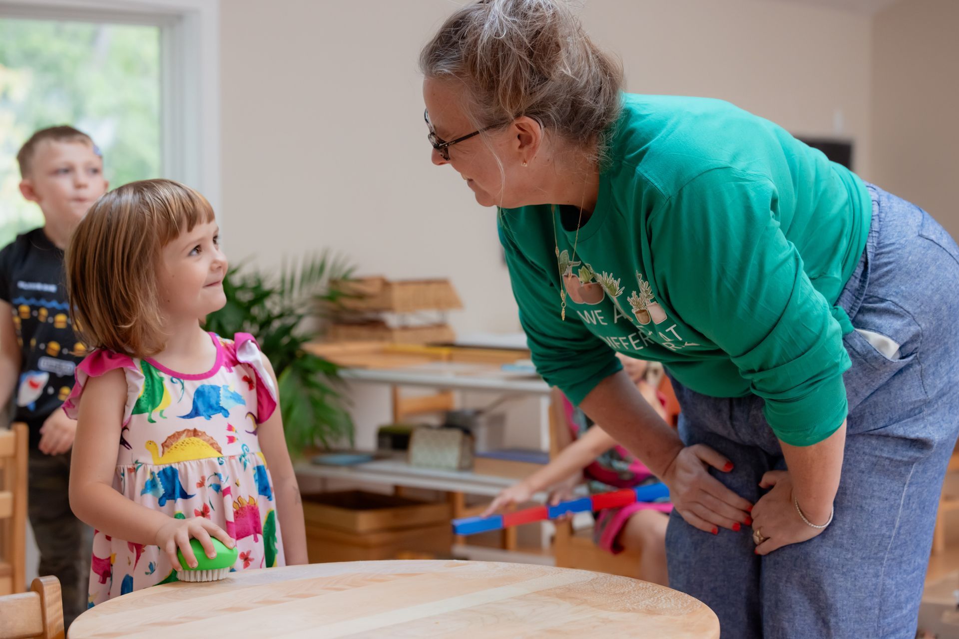 Teacher leaning toward a child at a classroom table while another child looks on.