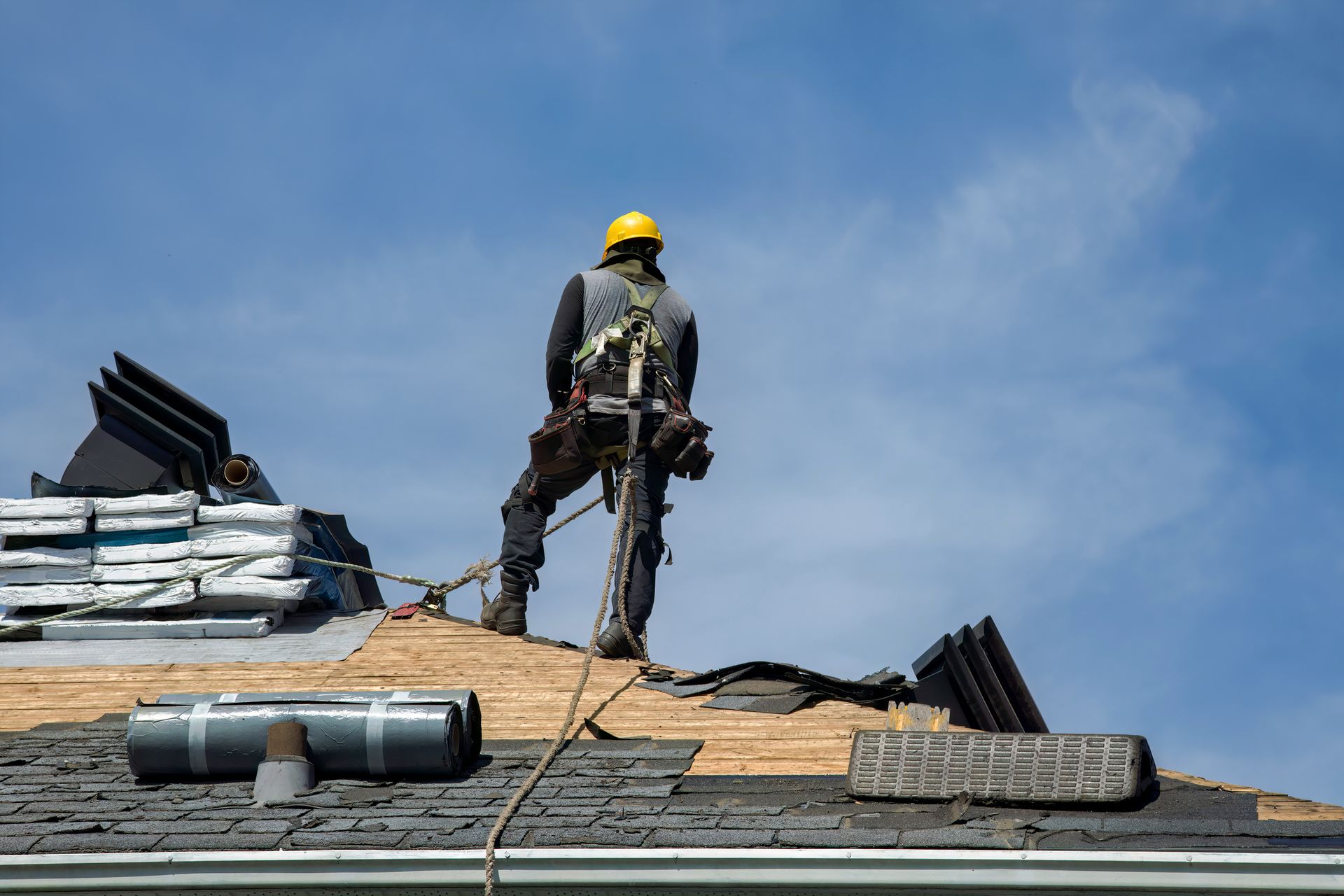 Roofer wearing safety harness stands on a roof, working on a sunny day.