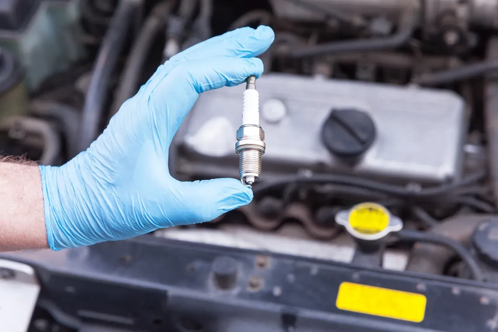 A person wearing blue gloves is holding a spark plug in front of a car engine.