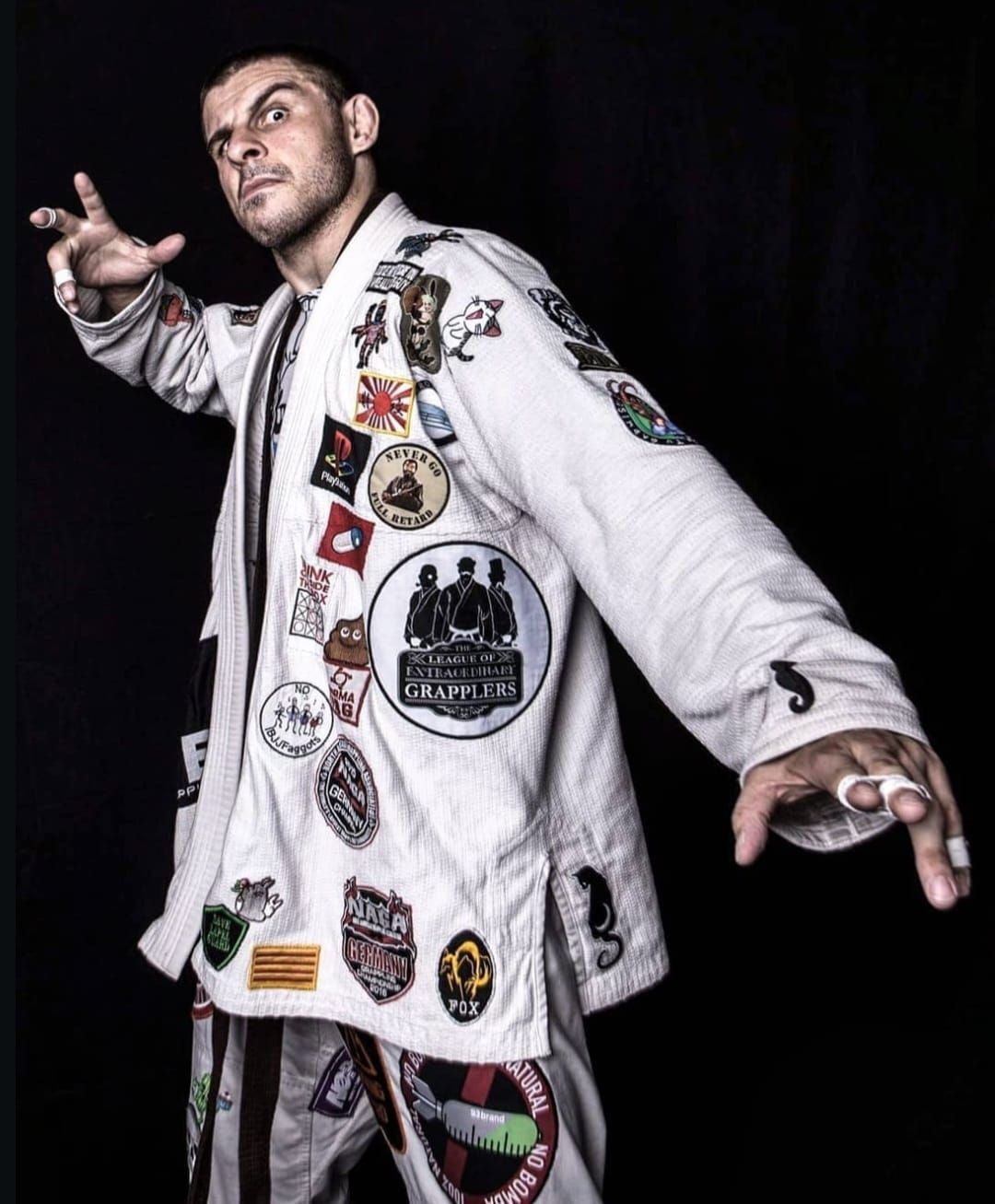 Mann in verziertem weißen Jiu-Jitsu-Gi, dunkler Hintergrund, gestikuliert mit den Händen, Aufnäher auf dem Gi.