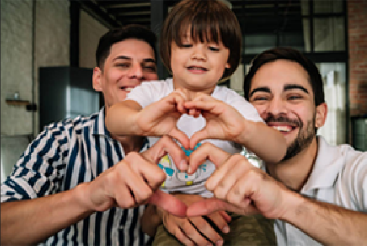 Two people smile as they hold a young child who is making heart shapes with their hands in an indoor setting.