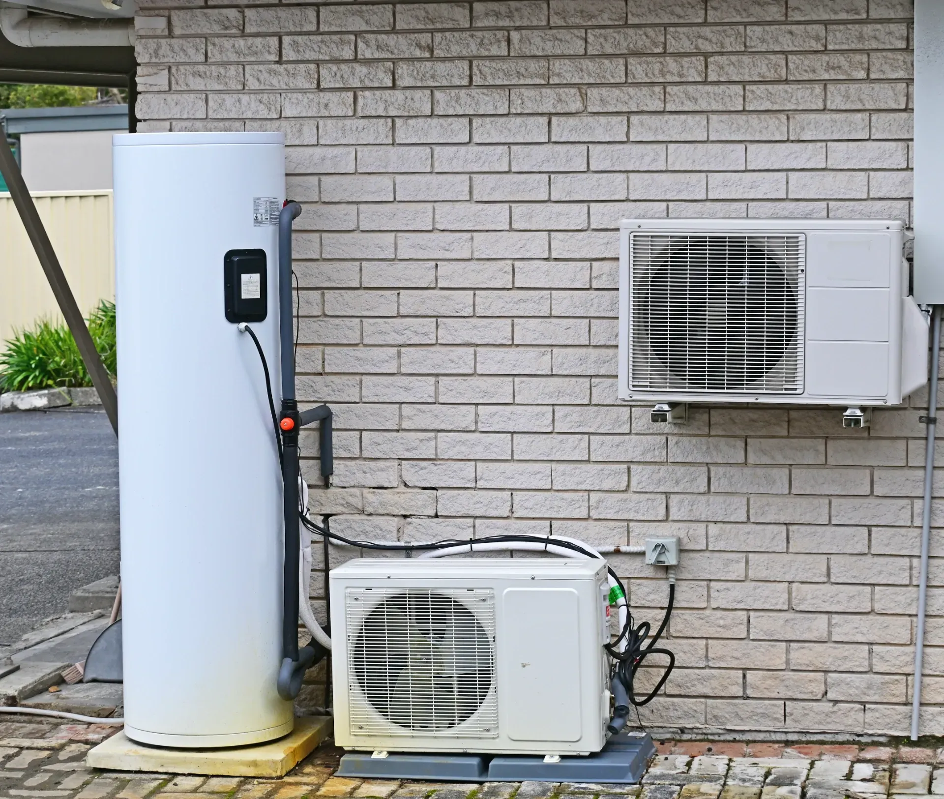 Outdoor heat pump and water heater units mounted beside a brick house wall. — Anthony Cameron Electrical & Solar In Woodgate, QLD