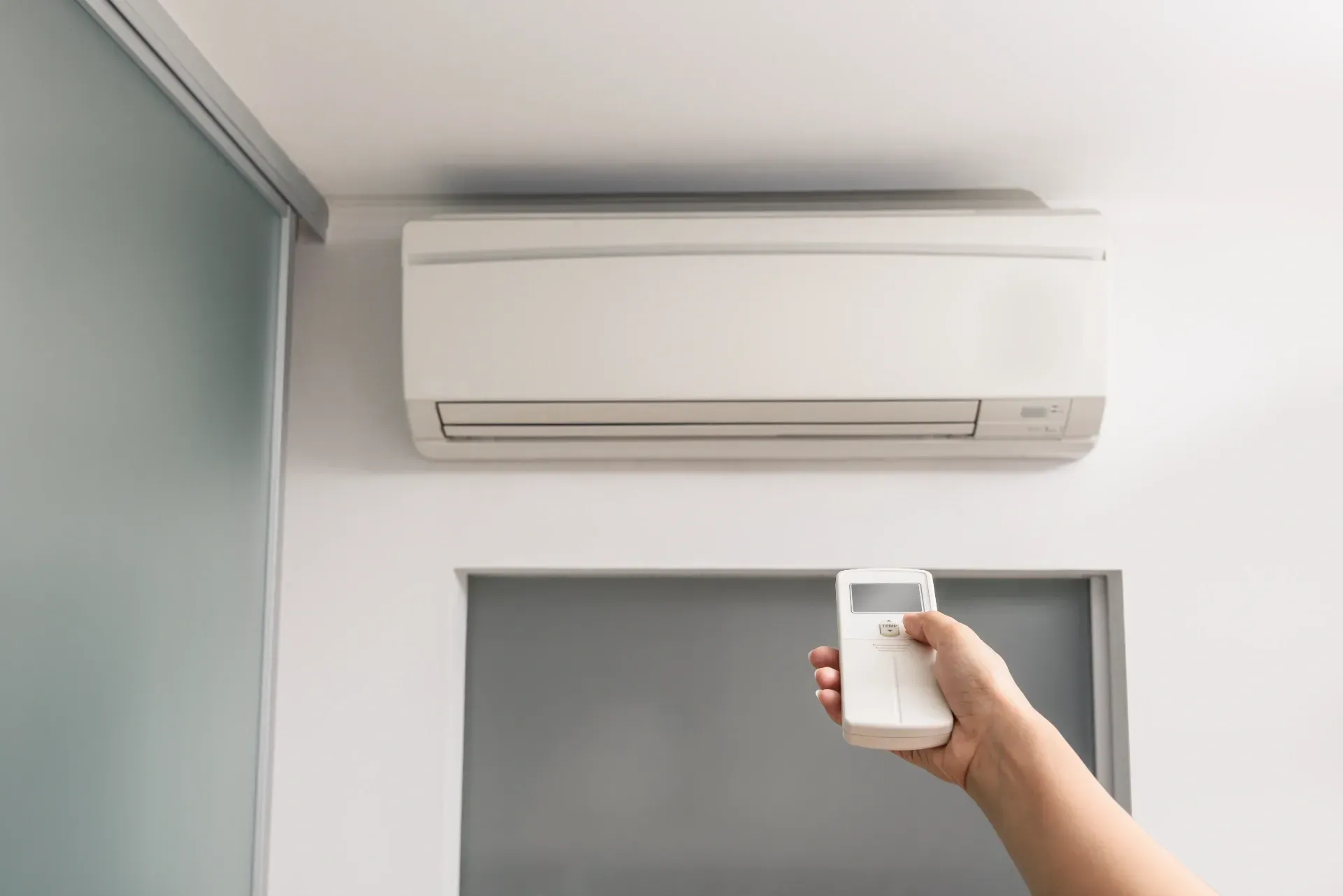 Hand holding a remote pointed at a wall-mounted air conditioner in a bright room — Anthony Cameron Electrical & Solar In Gin Gin, QLD