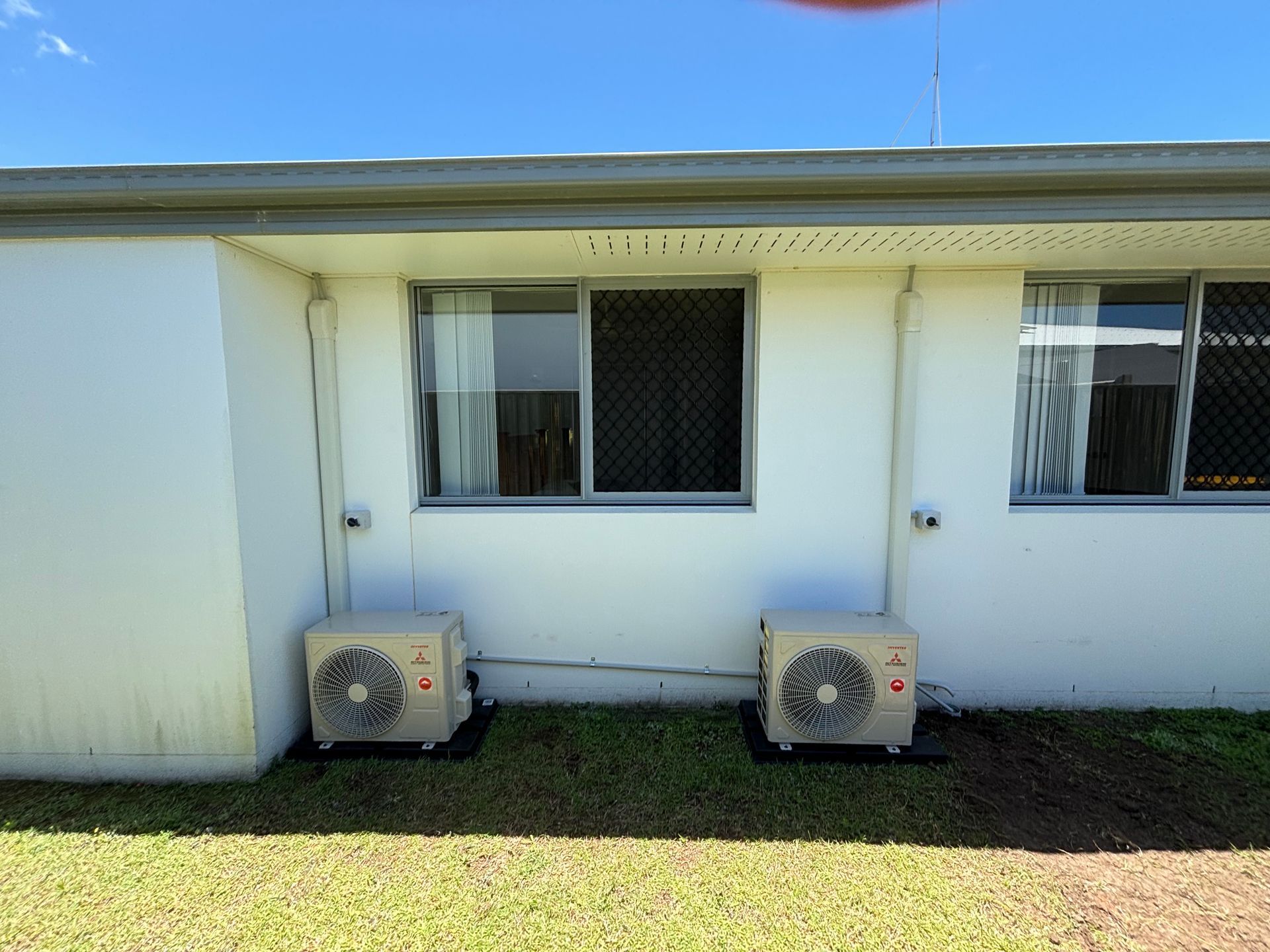 White house exterior with two air-conditioning units beneath windows on a grassy lawn — Anthony Cameron Electrical & Solar In Kepnock, QLD