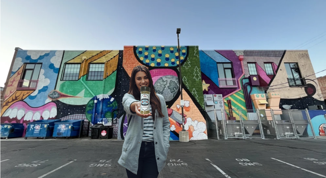 Woman in front of a colorful mural, holding an object toward the camera. Outdoor setting, blue sky.
