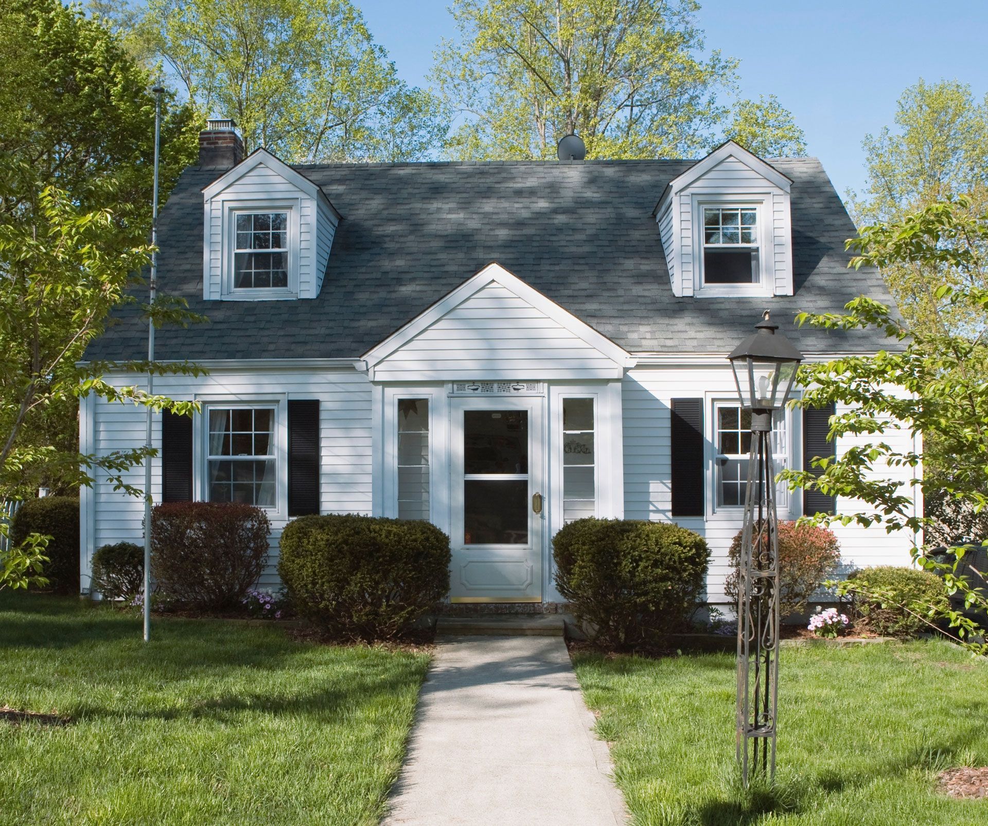 White house with dormer windows, front porch, and trimmed hedges beside a walkway under leafy trees