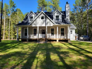 A two-story, grey, single-family home with a front porch, dormer windows, and a tall chimney, situated in a wooded lot.