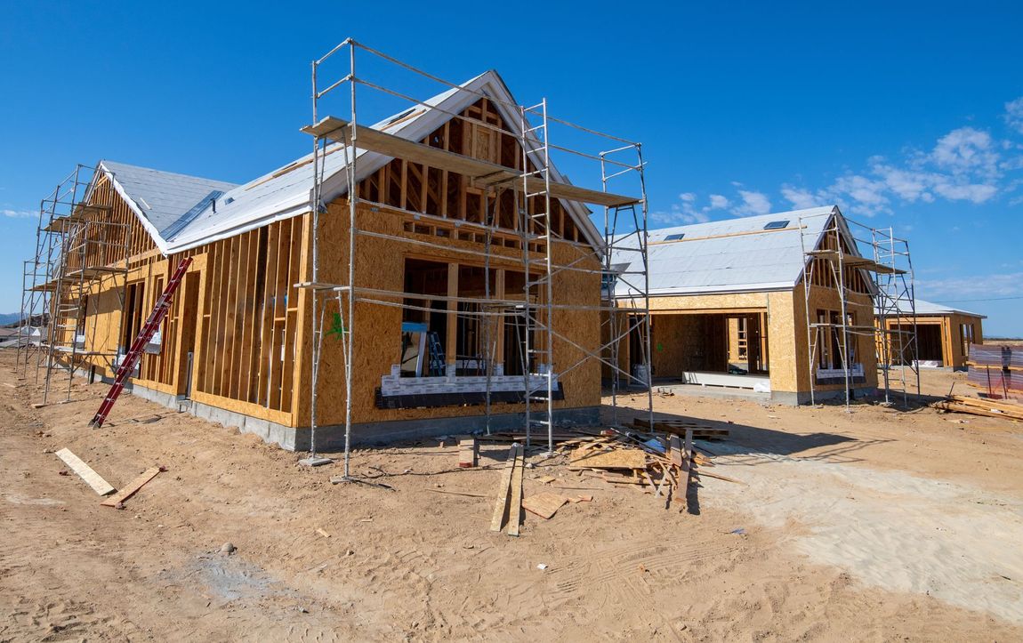 Wood-framed houses under construction; scaffolding, blue sky, and dirt ground.