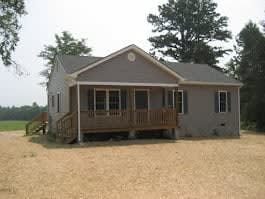 A gray, one-story house with a wooden porch and stairs, surrounded by a dry, grassy yard and trees.