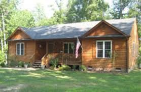 A single-story wooden cabin house with a covered front porch, grey shingled roof, and green lawn surrounded by trees.