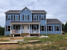 Blue two-story house with white porch and dark shutters, set on a grassy lot under a cloudy sky.