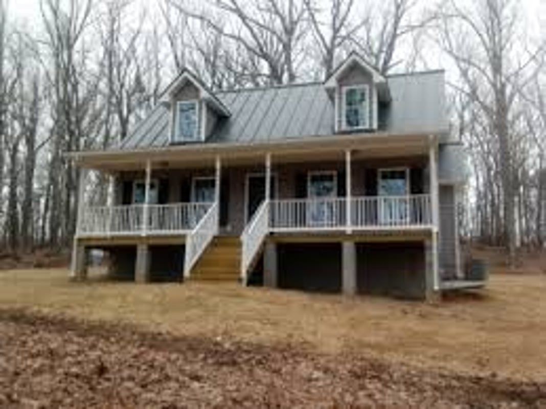 A tan house with a metal roof and a raised front porch sits in a wooded area with dry grass in the foreground.