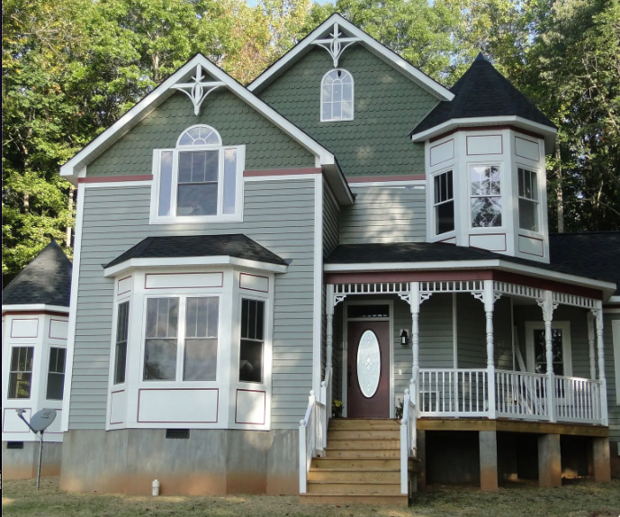 Victorian-style green house with white trim, wraparound porch, and bay windows in a wooded setting