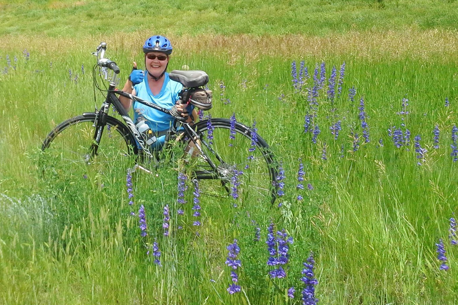 Woman on bicycle in tall green grass, smiling, surrounded by purple wildflowers.