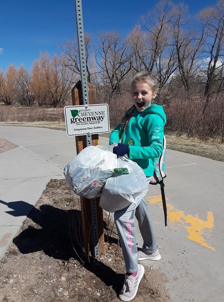 Girl smiling, holding two full trash bags near a trail sign that reads