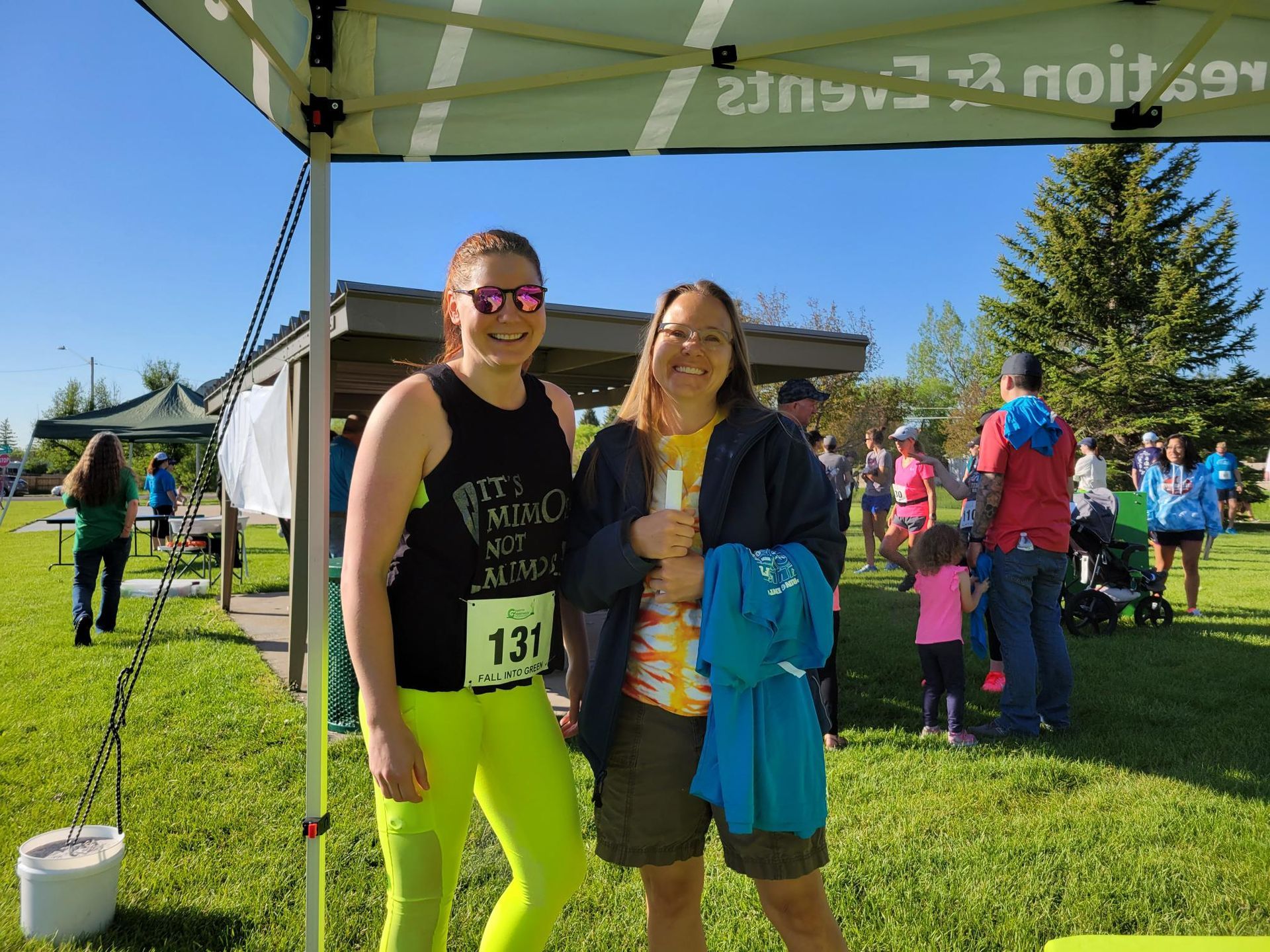 Two smiling women pose at a park event. One wears neon workout clothes, the other holds a blue shirt.