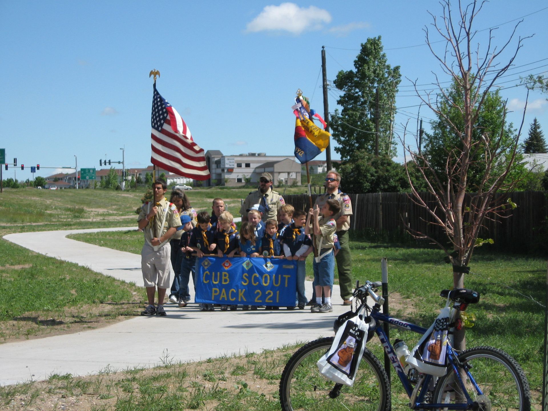 Cub Scout Pack 221 holds flags on a walking path with a blue banner. American flag waves in the background.
