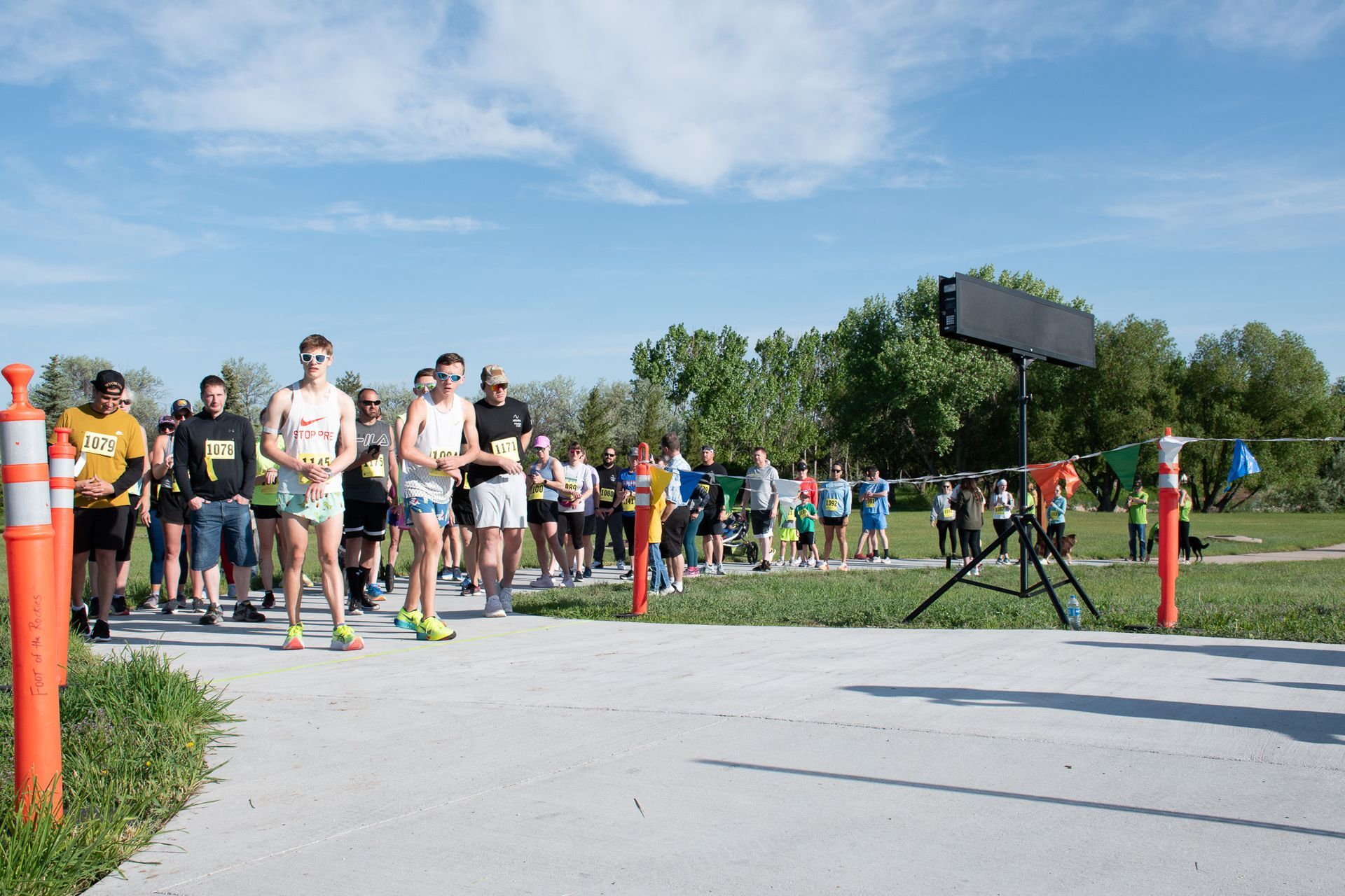 People lined up at a running event, ready to start on a paved path, sunny day.