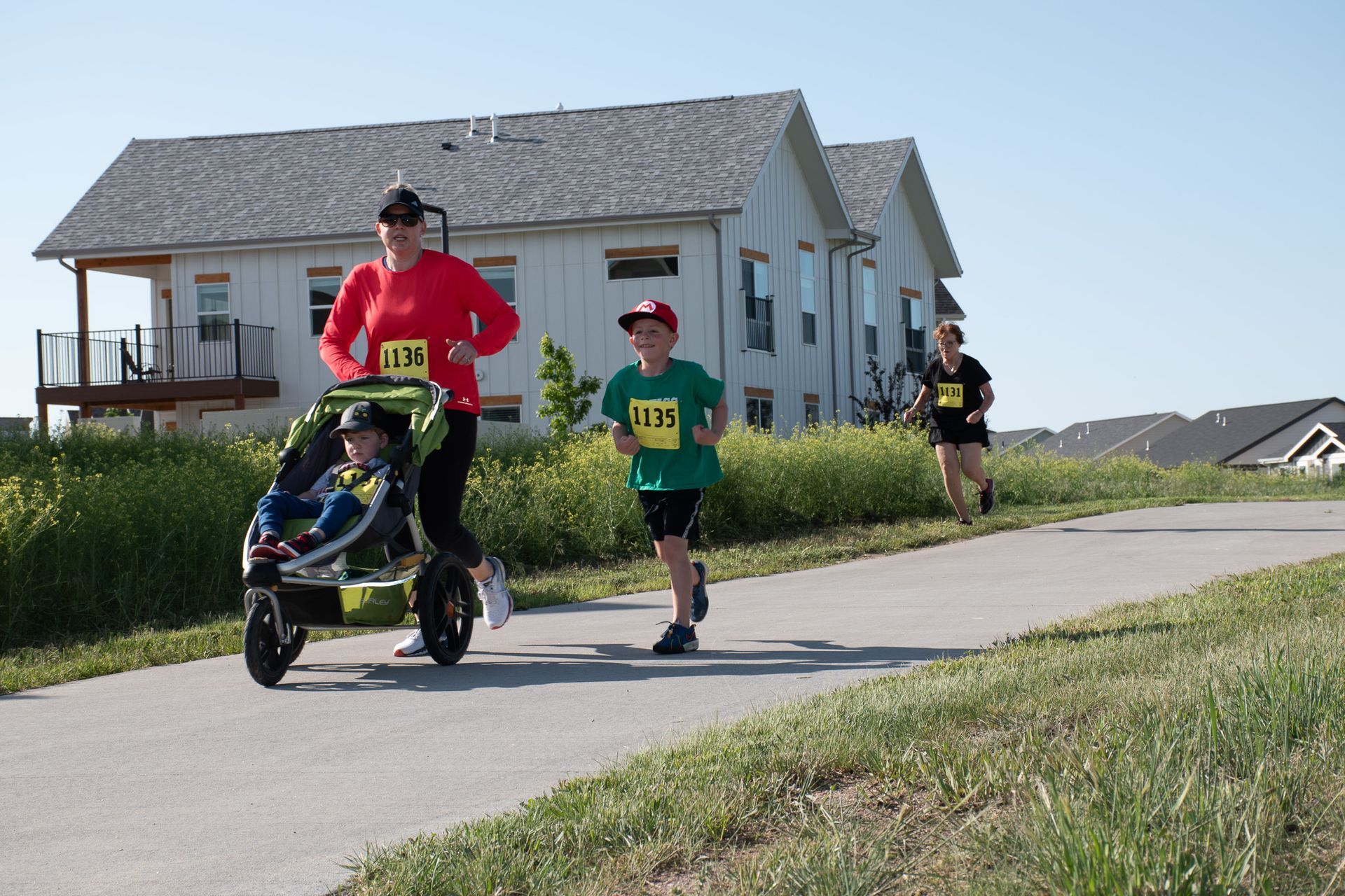 People running on a paved path in a race. A runner pushes a stroller. A child runs nearby. Buildings in the background.