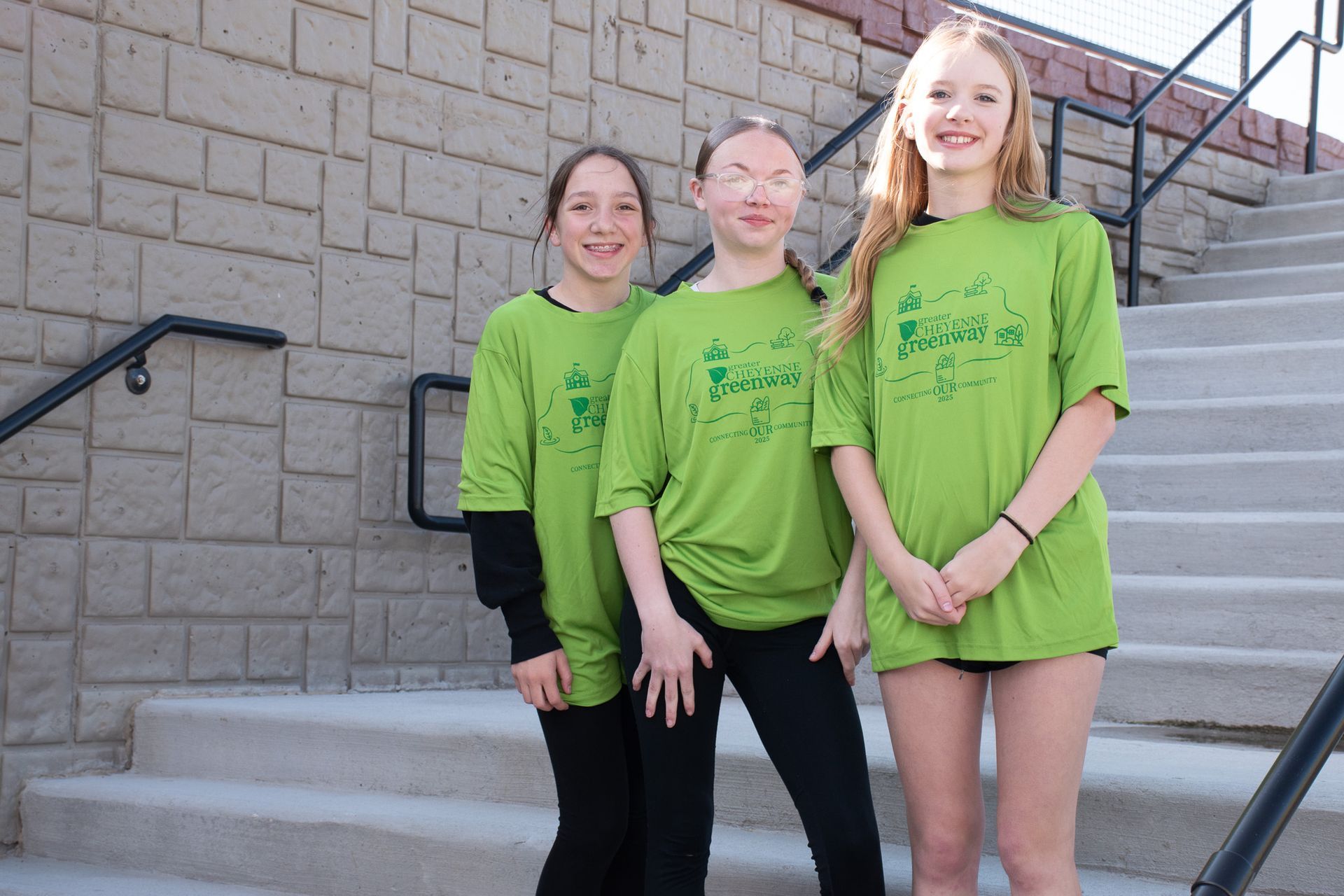 Three people in green shirts stand on steps, smiling. Brick wall and black railing in background.