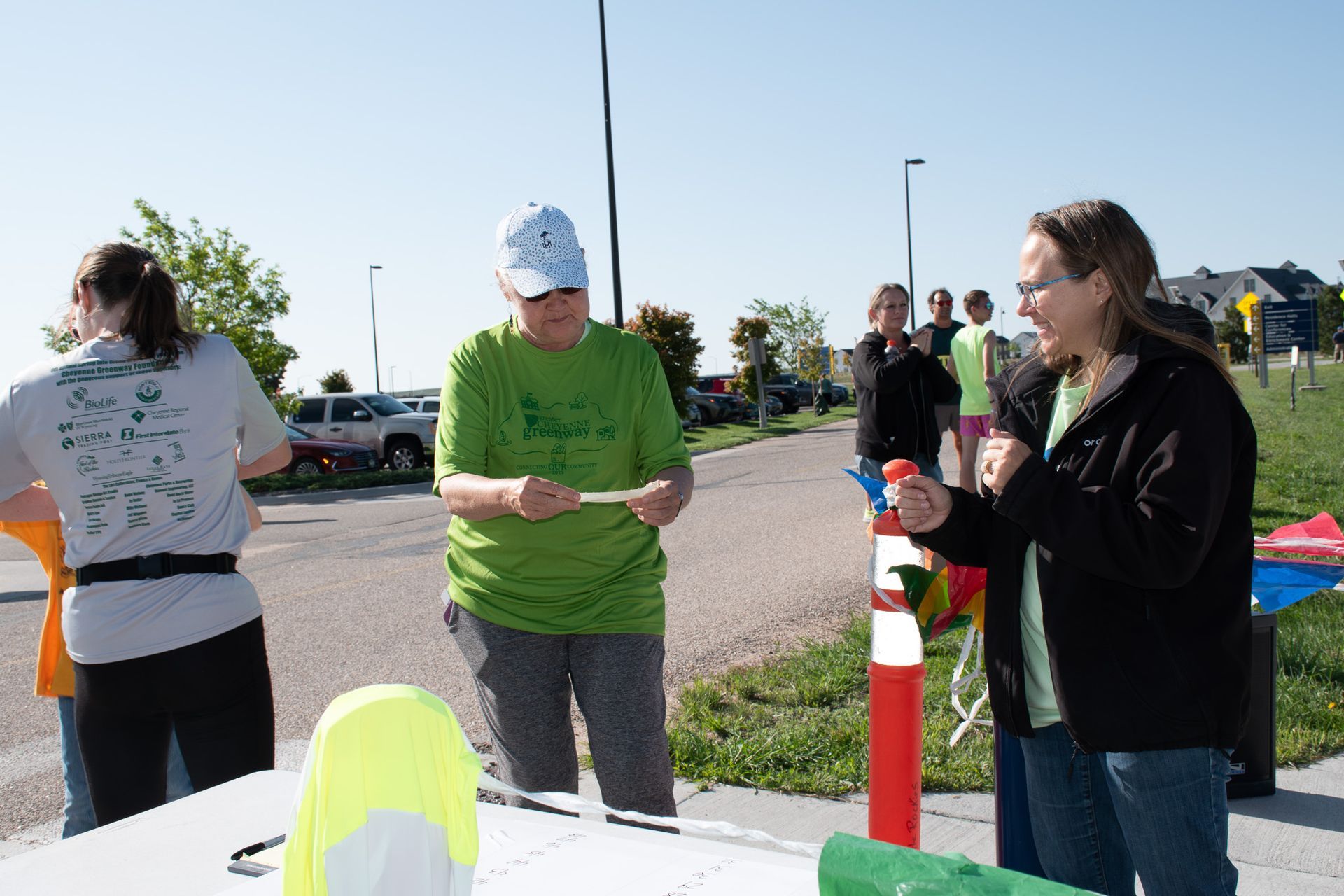 People at a table outdoors, one holds a paper, another with a red item. Paved area, bright day.