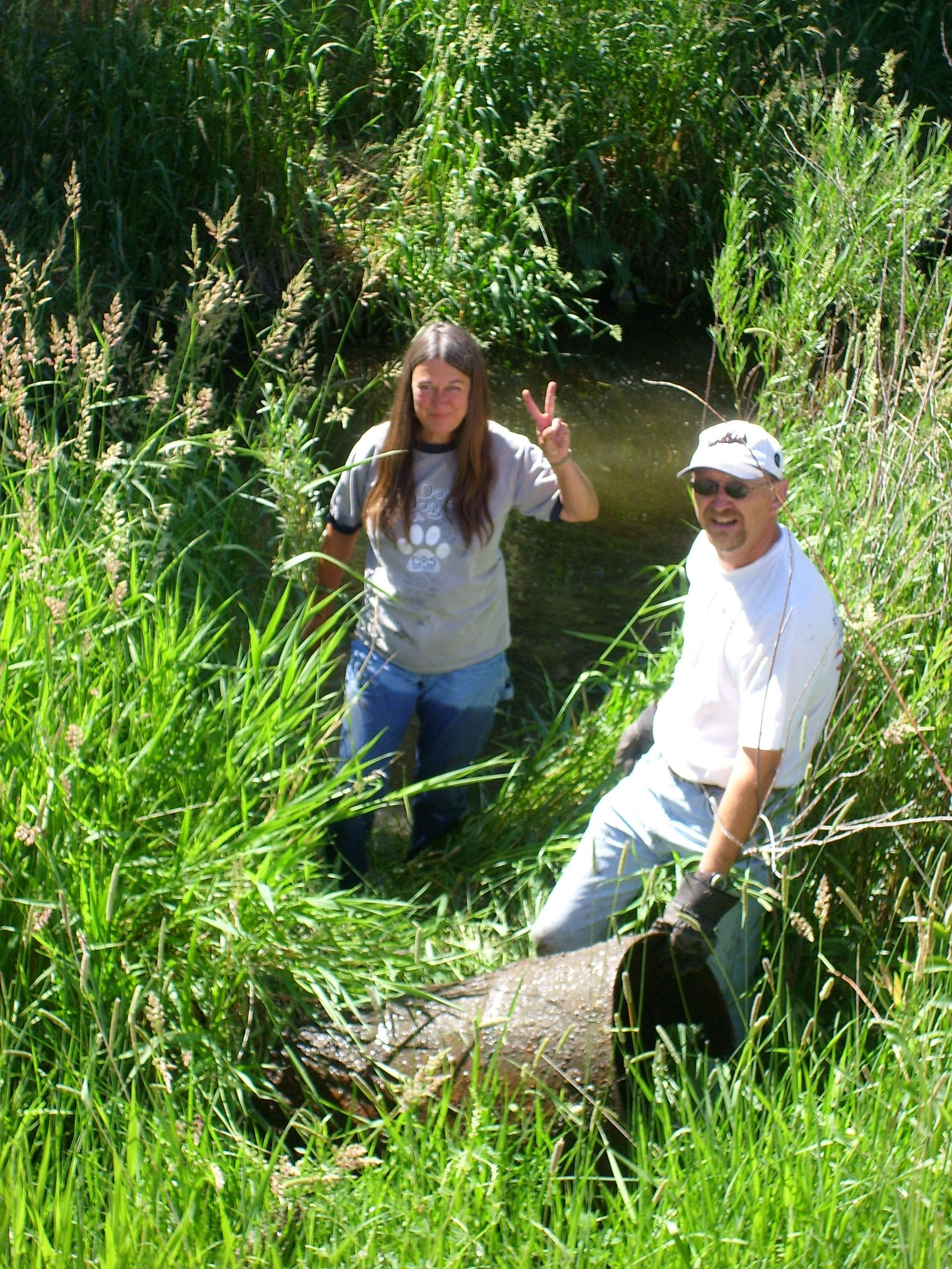 Two people by a stream. One makes peace sign, the other smiles. Both are in grassy area.