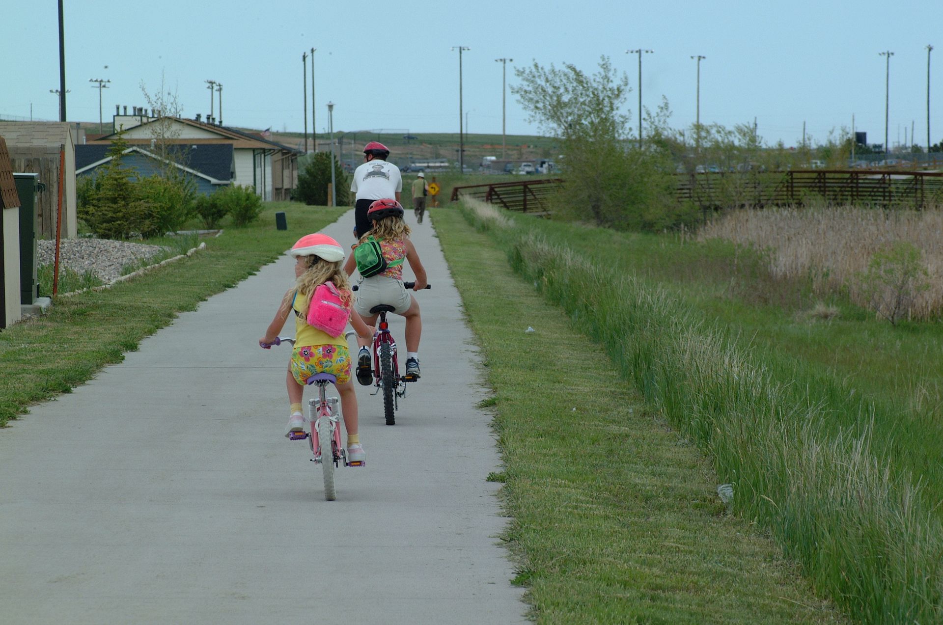 Three children riding bikes on a paved path next to a grassy area, wearing helmets.