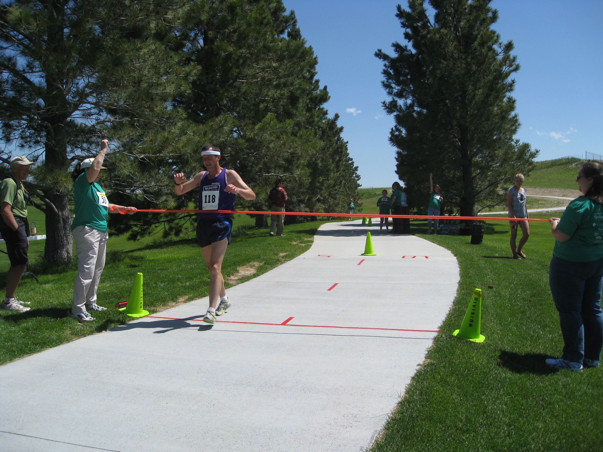Runner crossing finish line on a paved path, cheered on by spectators. Sunny, outdoor setting.