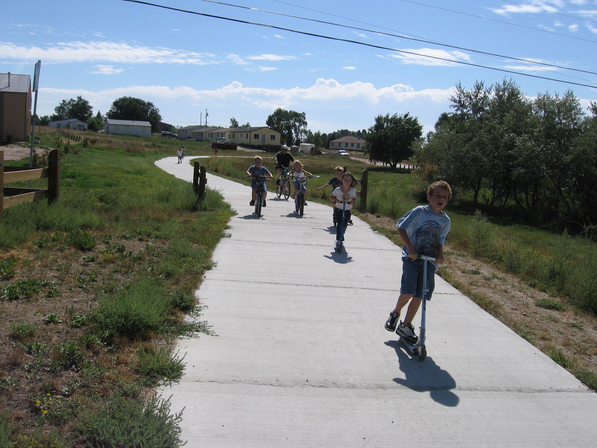 Children scootering on a paved path outdoors. Sunny day, green grass, buildings in the background.