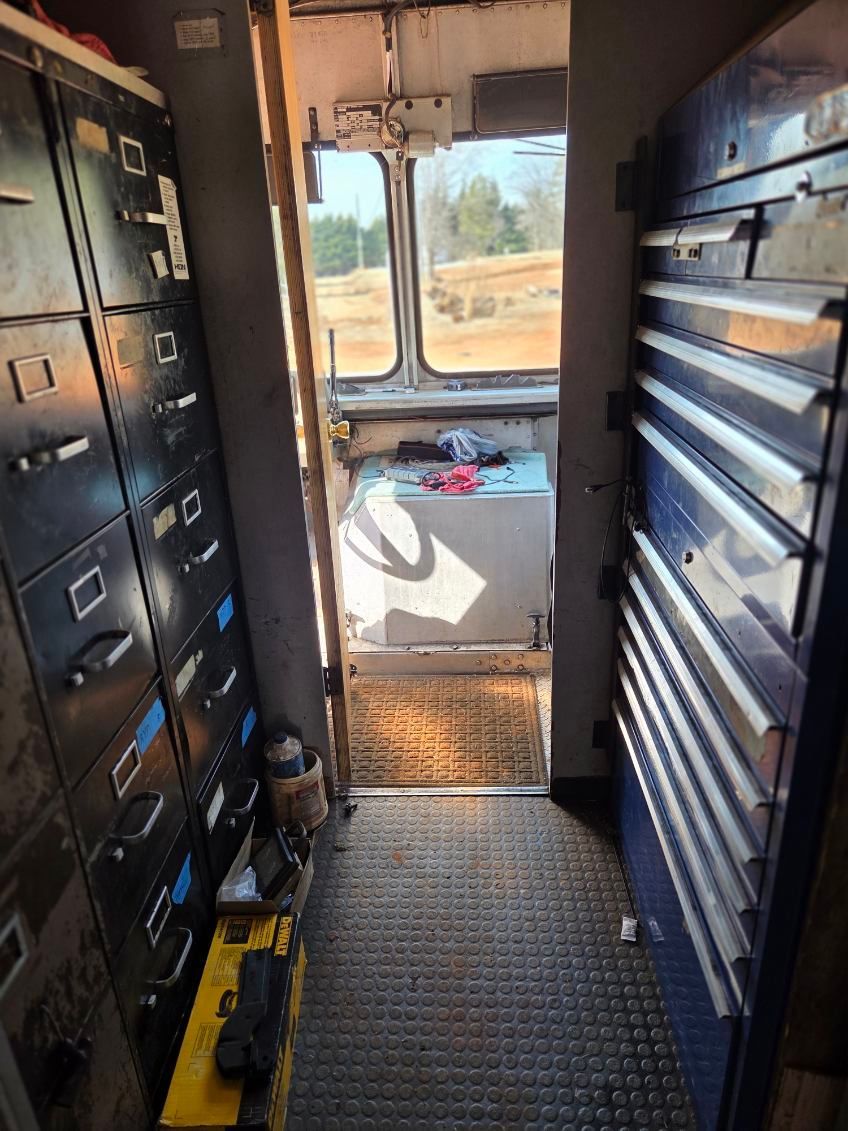 Inside a narrow vehicle interior with metal shelving, a rear door open to a bright outdoor scene.